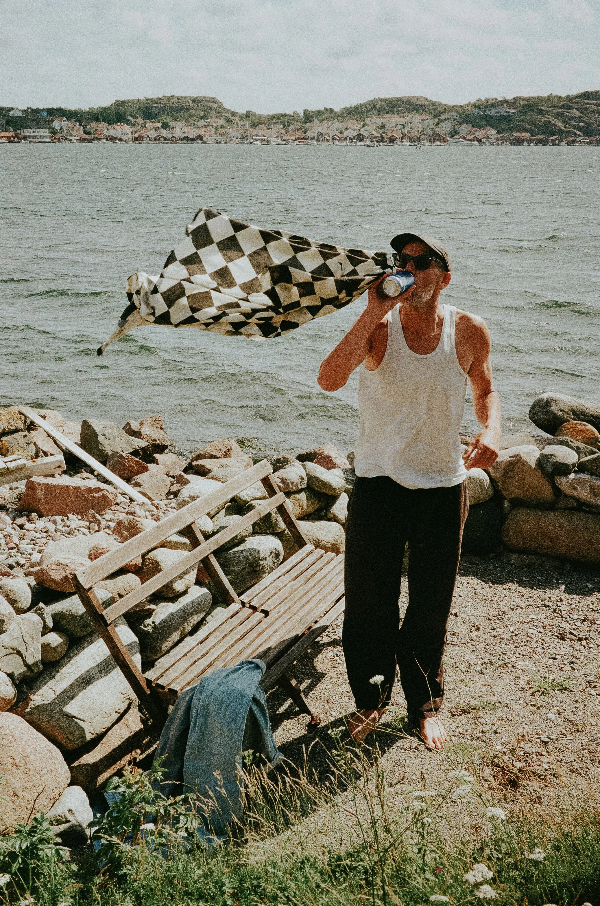 A man standing barefoot on a rocky beach, wearing a white tank top, black pants, sunglasses, and a cap, holding a checkered umbrella and drinking from a can. There is a wooden bench with a denim jacket draped over it and rocks lining the shoreline, w