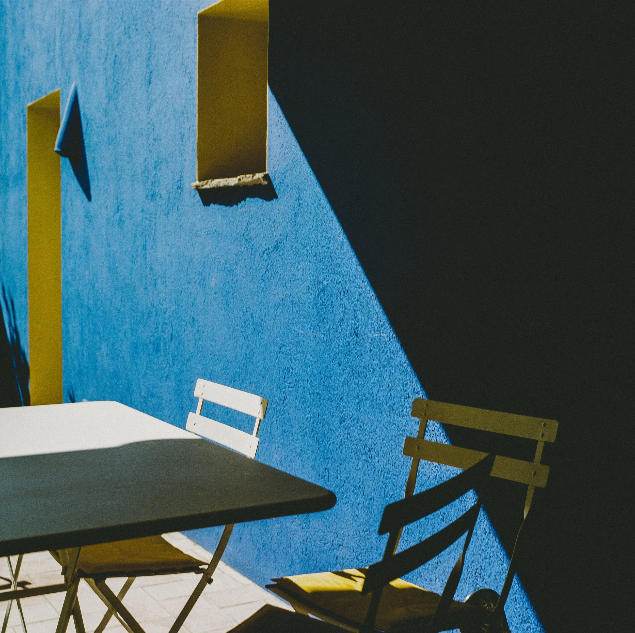 Outdoor seating area with yellow and white chairs, a black table, a bright blue wall with a small square window, and shadows cast by sunlight.