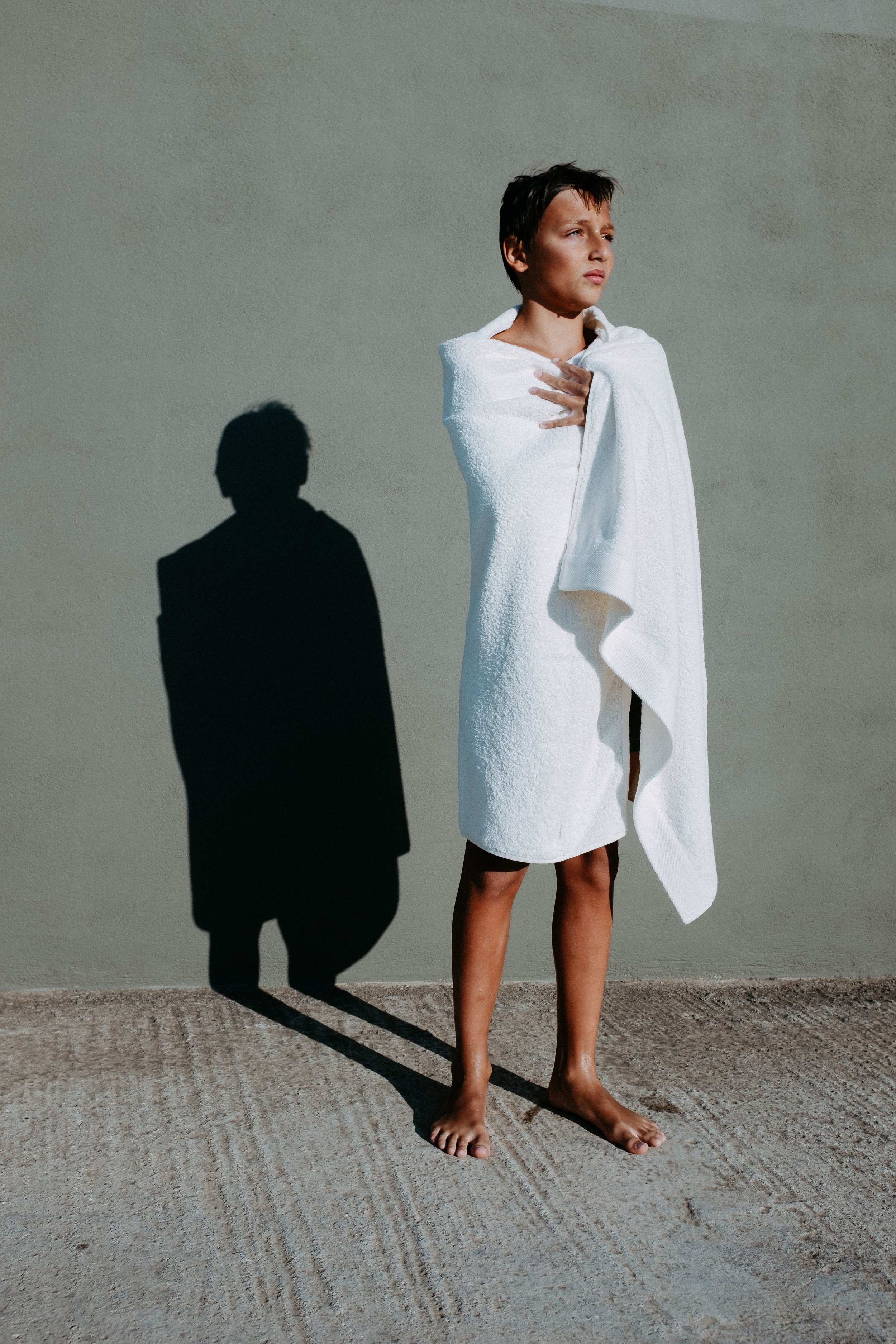 A person with short dark hair standing barefoot on a textured floor against a plain wall, wrapped in a white towel with a contemplative expression, casting a distinct shadow on the wall.
