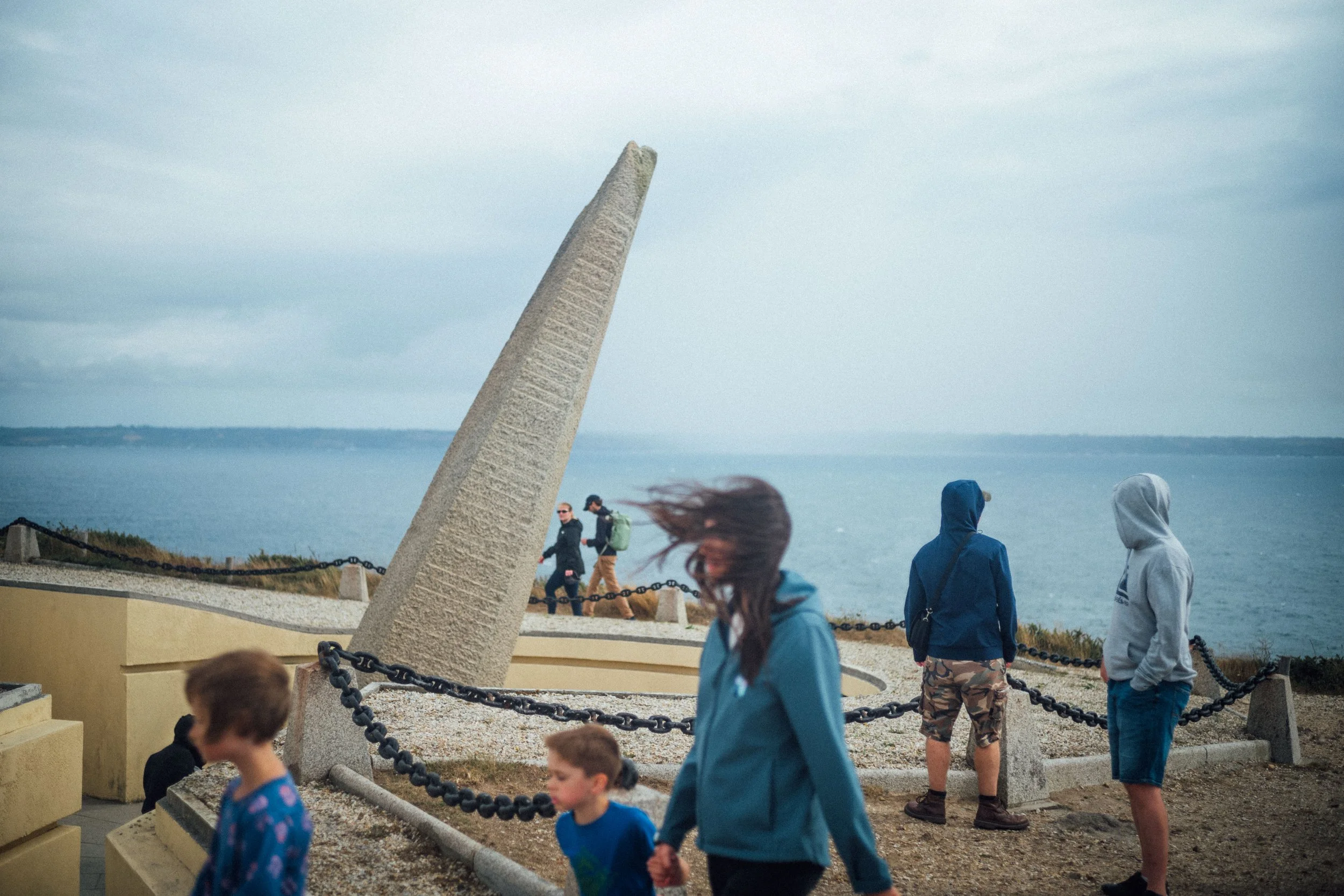 People walking near a seaside sculpture with a large inclined stone structure, on a cloudy day.