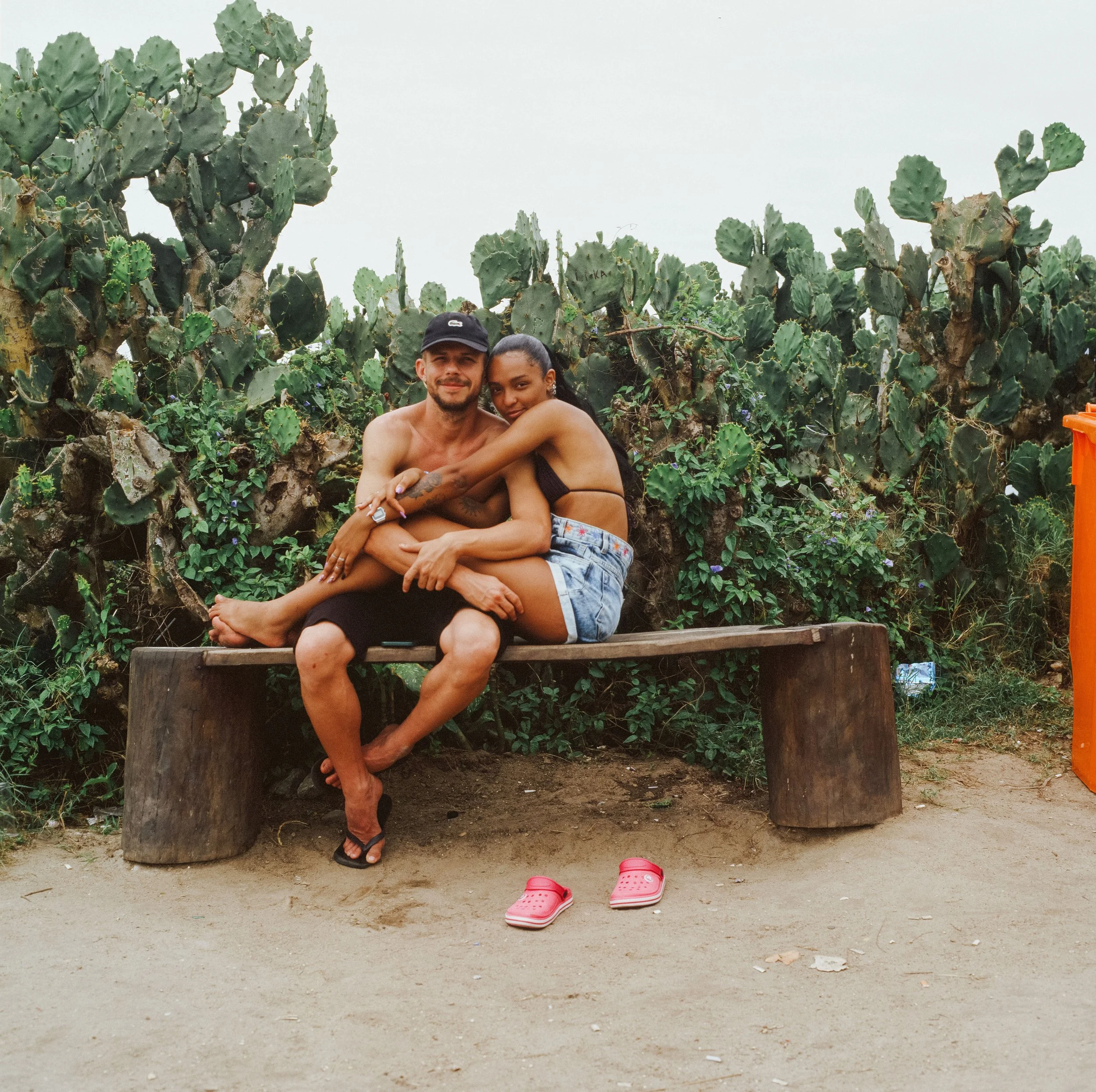 A couple sitting on a wooden bench in front of cactus plants, embracing and smiling at the camera, with pink sandals on the ground in front of them, and an orange trash bin to the right.