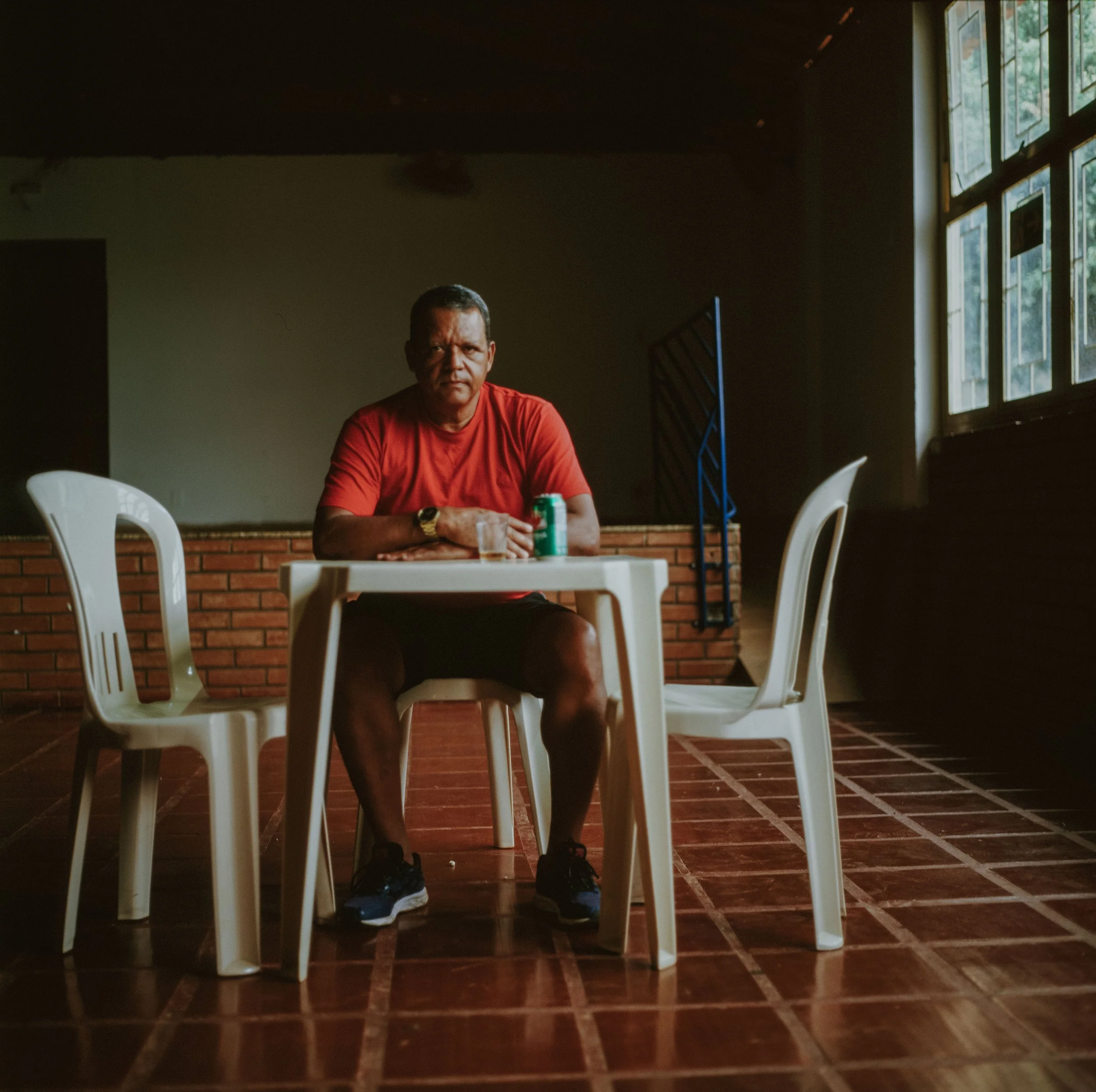 A man sitting at a white plastic table with two empty chairs, in a dimly lit room with large windows on the right, a brick wall behind, wearing a red shirt and black shorts, with a glass and a soda can on the table.
