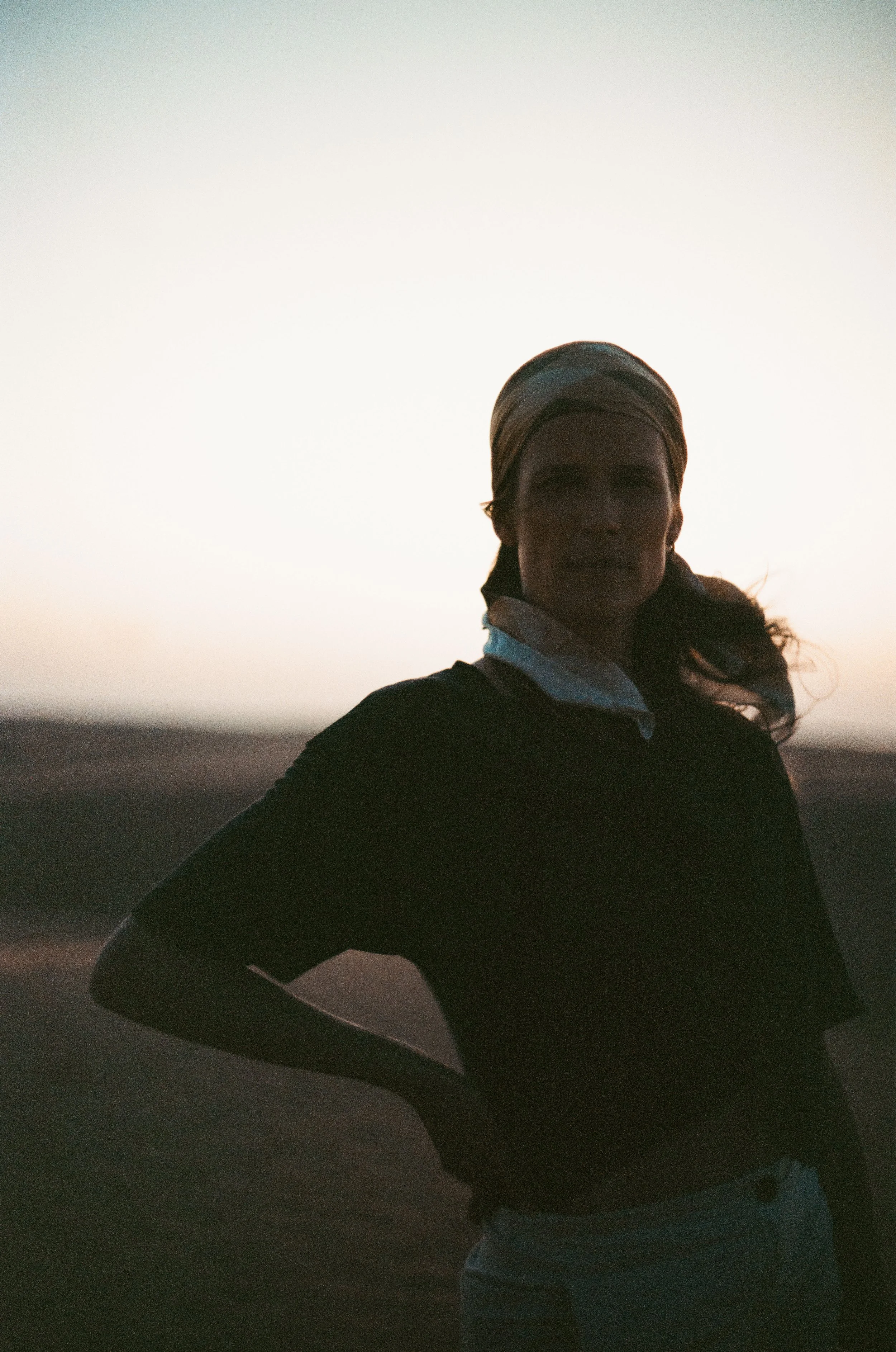 Silhouette of a woman standing outdoors during sunset or sunrise, wearing a headscarf, black top, and jeans, with a neutral background.