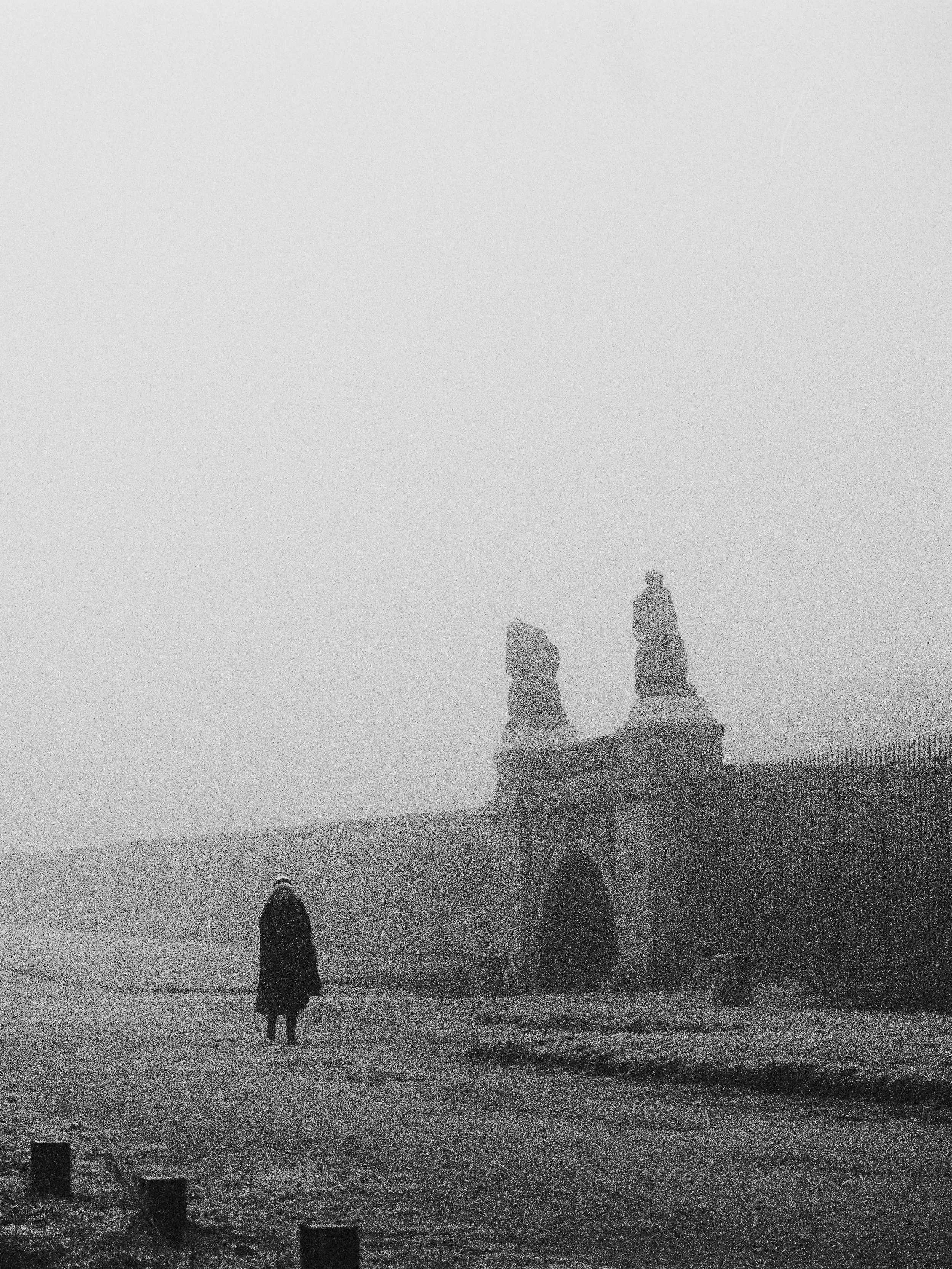 A person walking alone on a foggy, deserted beach near a historic stone archway with statues on top.