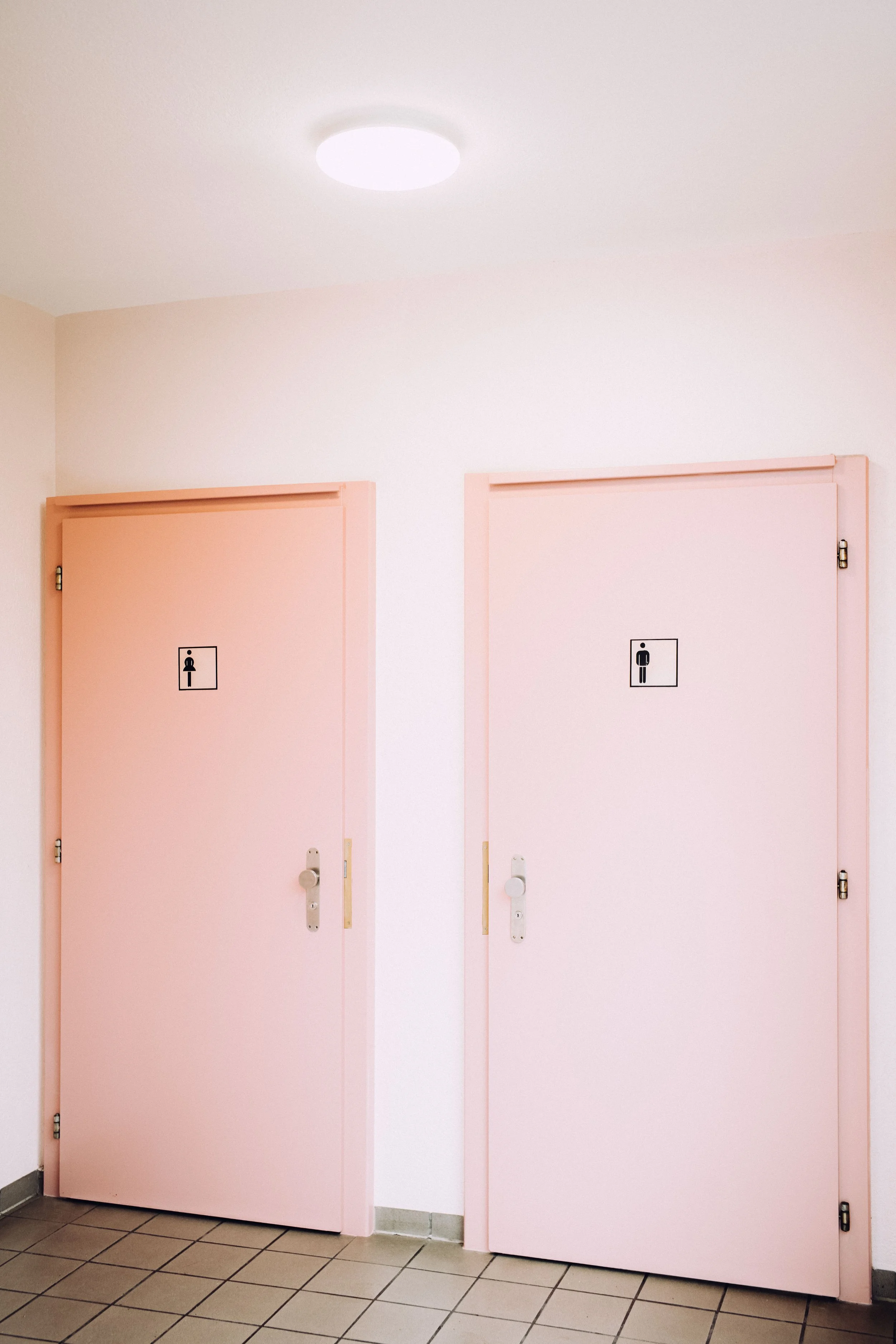 Pink doors with men's and women's restroom signs, located in a tiled hallway under an overhead light.