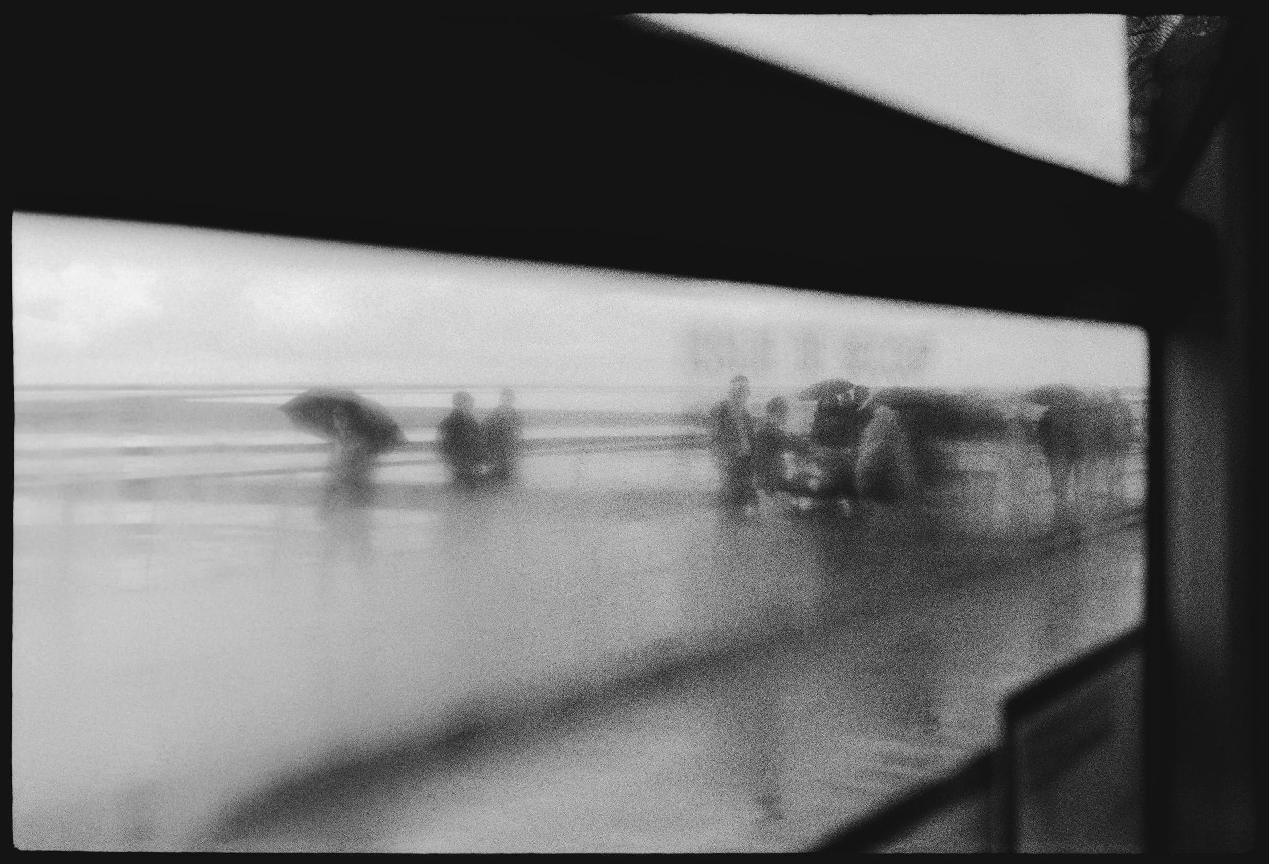 Blurred black and white photo of people with umbrellas walking on a rainy bridge, viewed through a bus window.