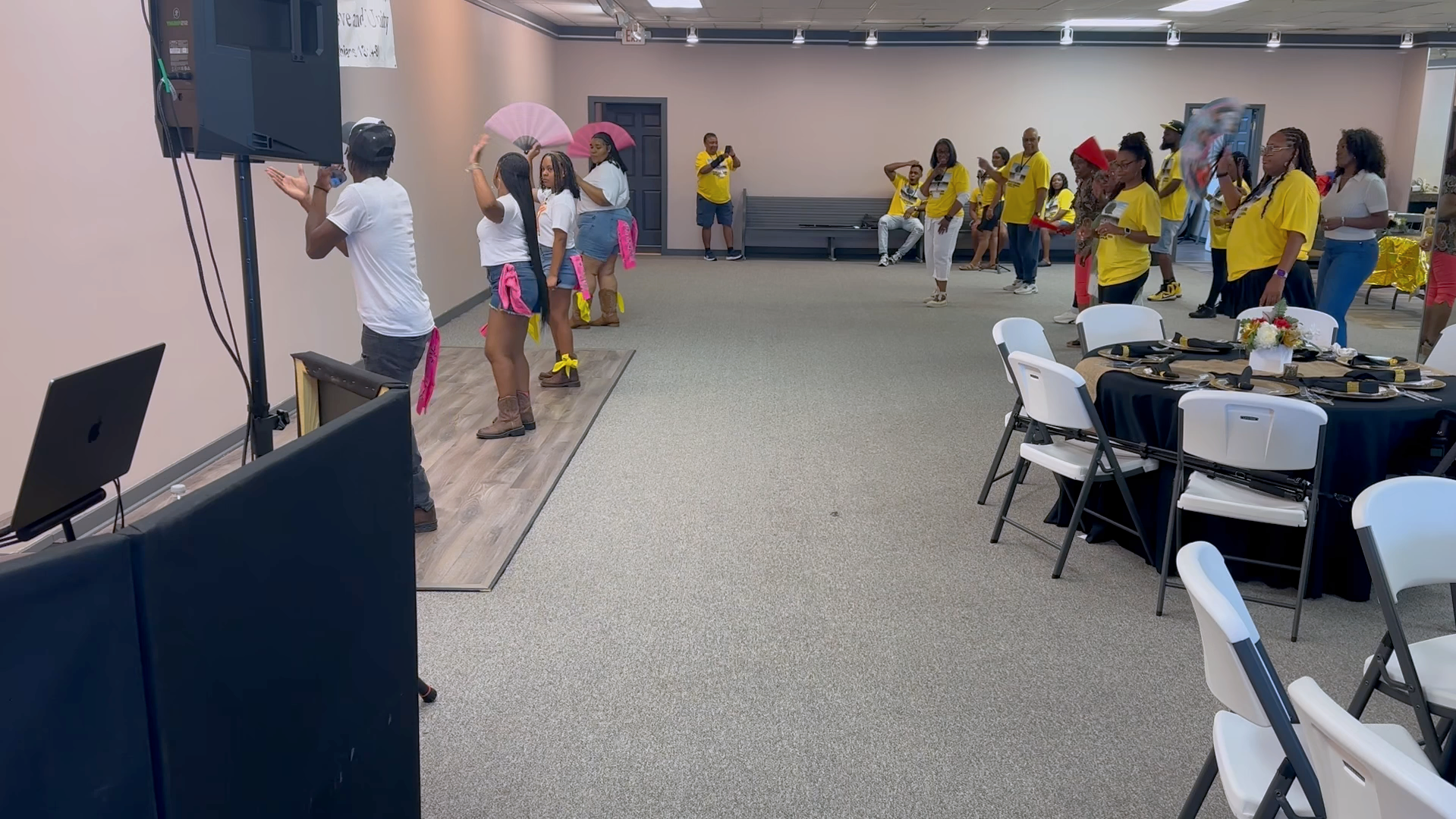 People dancing and socializing at an indoor event with decorated tables. Some individuals are in white shirts, others in yellow shirts. A person is taking a photo, and some are holding umbrellas. The setting appears to be a banquet or celebration.