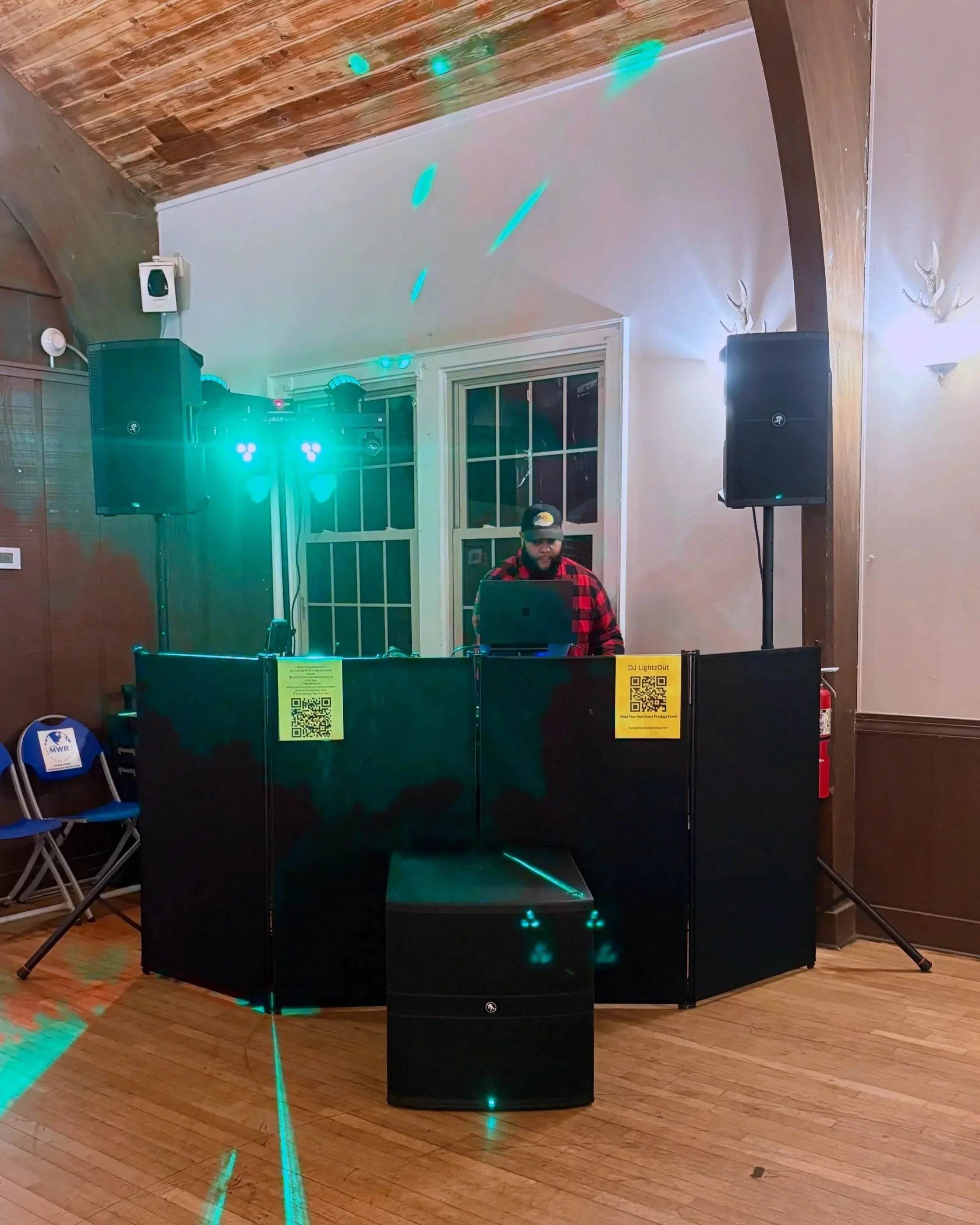 A man DJing at a party with teal lighting, two large speakers on stands, and QR codes on the DJ booth.