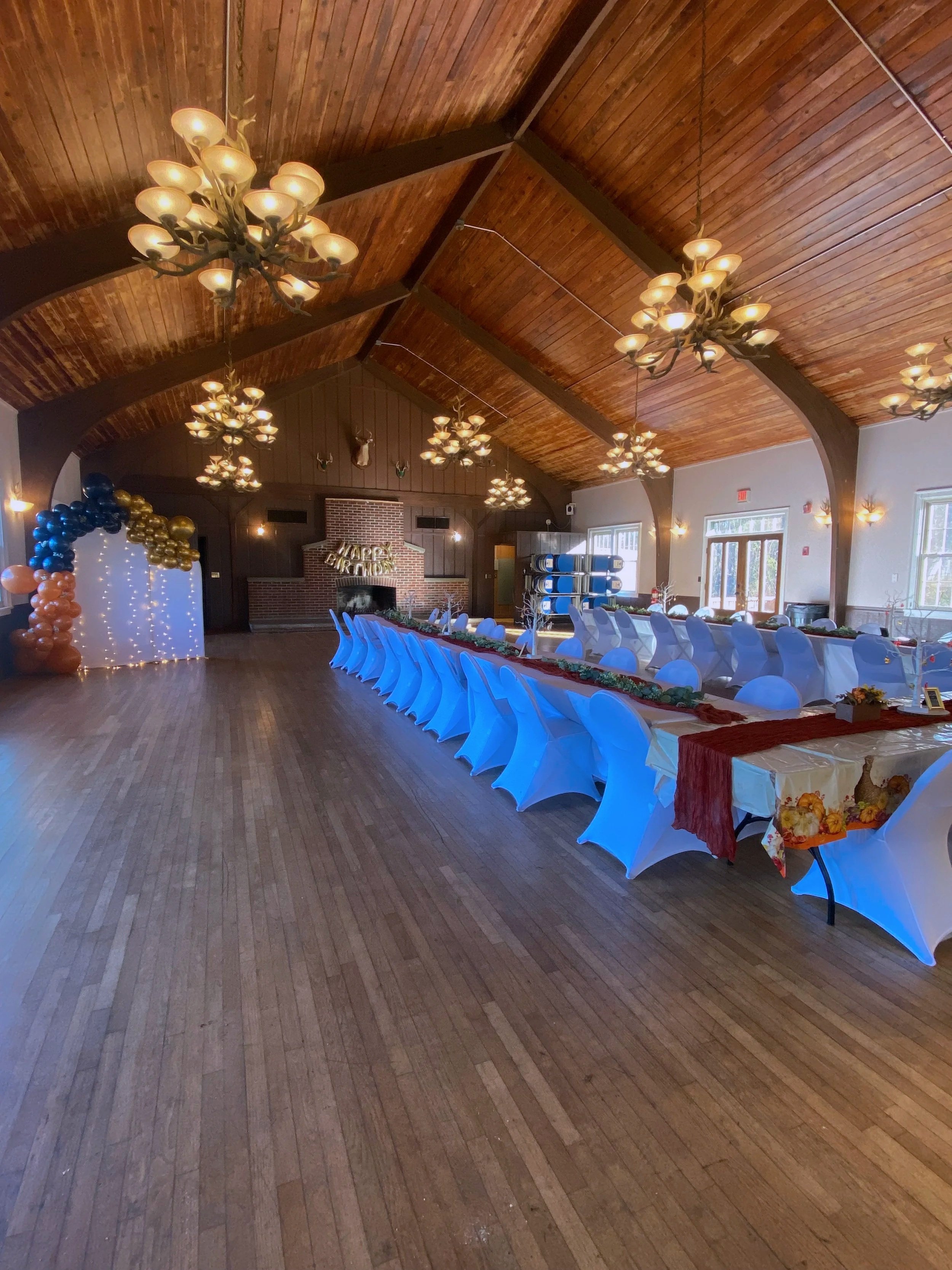 An elegantly decorated event space with long banquet tables covered in creamy tablecloths, adorned with red runners and autumn-themed centerpieces. White covered chairs surround the tables. The room has a vaulted wooden ceiling with multiple chandeliers, and there are large windows letting in natural light. Balloons and decorative lights are seen in the background, indicating a celebration or party.