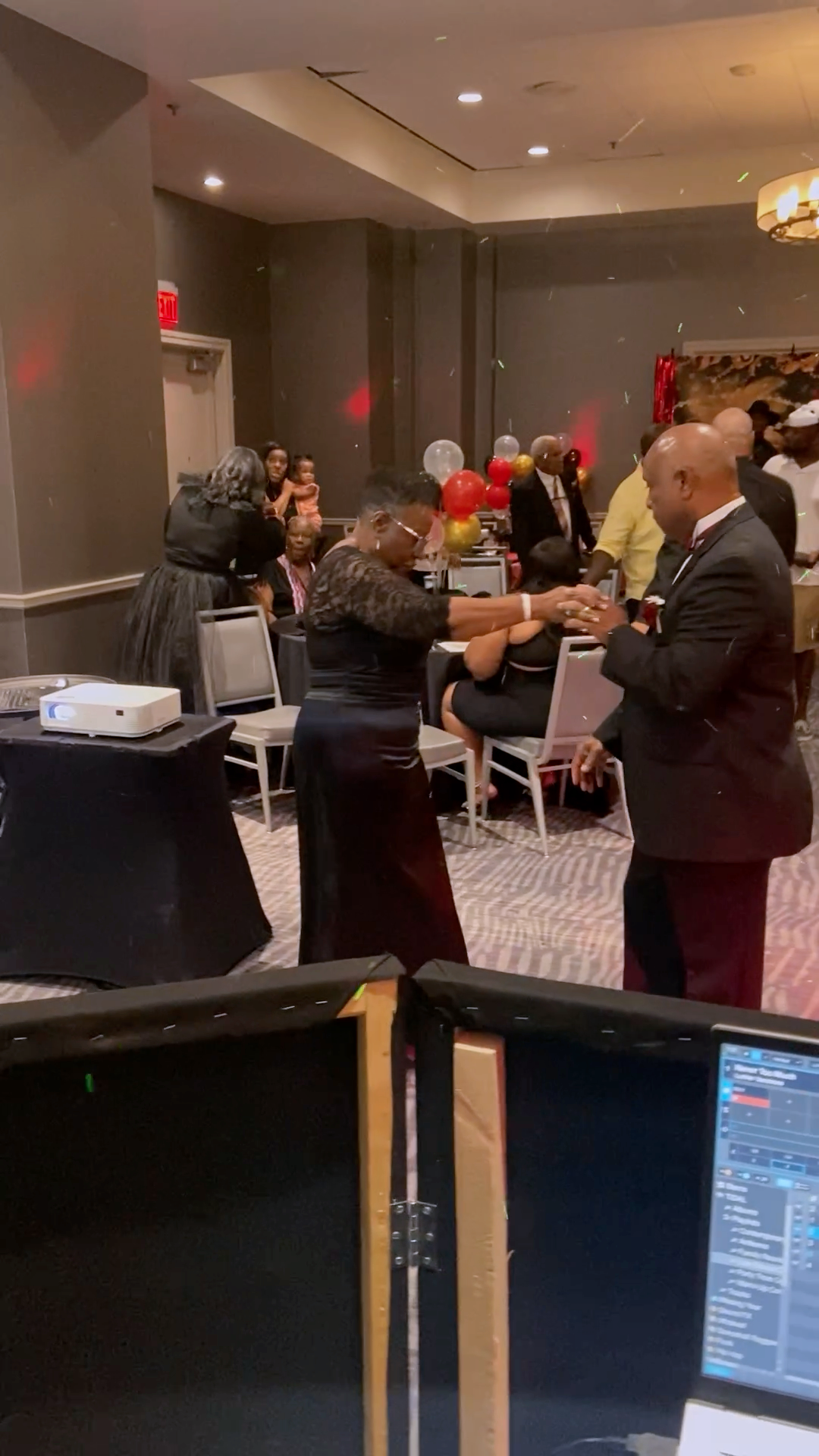A woman and a man are dancing together at a formal event, with other guests seated at tables decorated with balloons in the background.