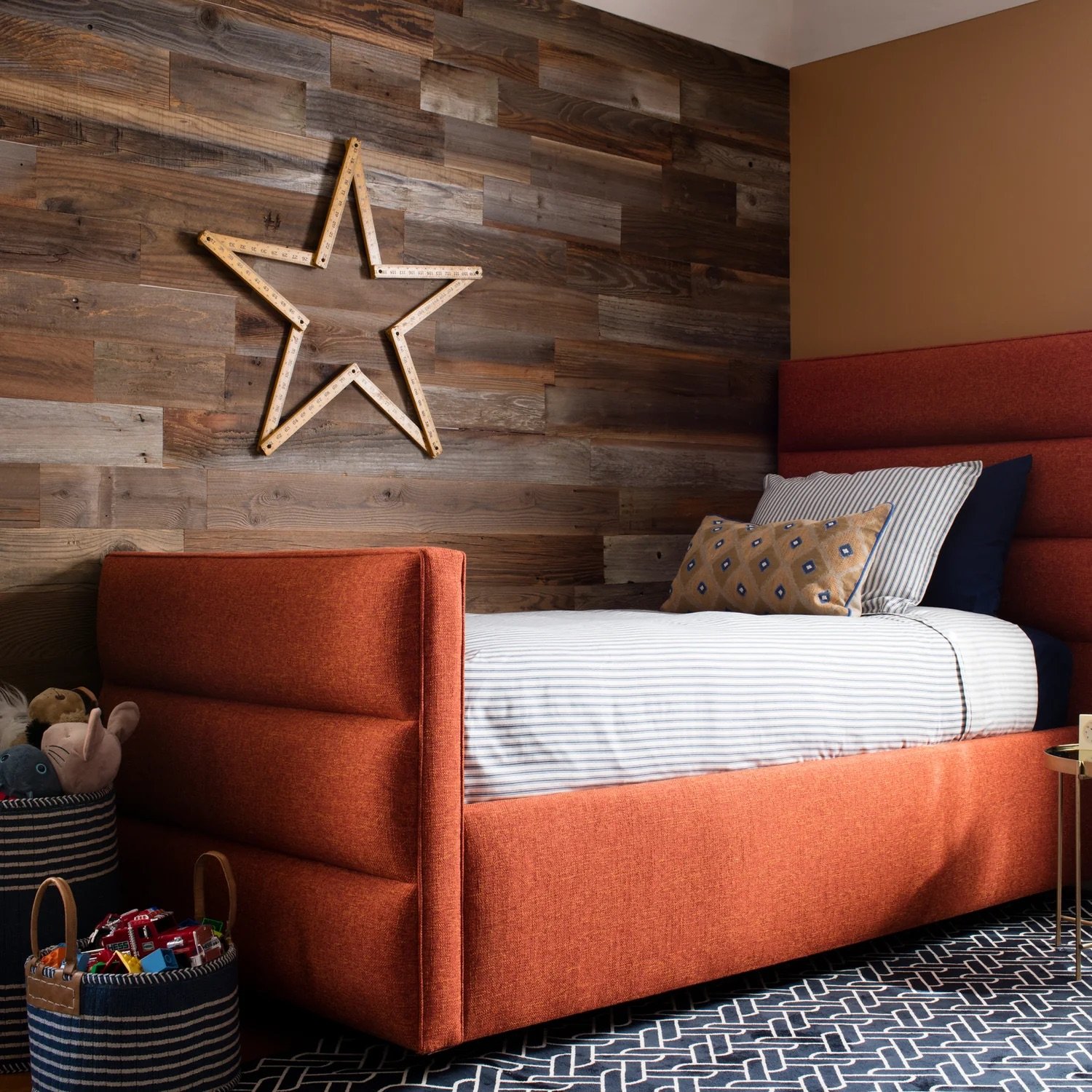 A cozy bedroom with a red upholstered platform bed, striped white and gray bedding, and various throw pillows. The wall behind the bed features a wooden plank design with a wooden star-shaped decor. There are baskets with plush toys and toys at the f