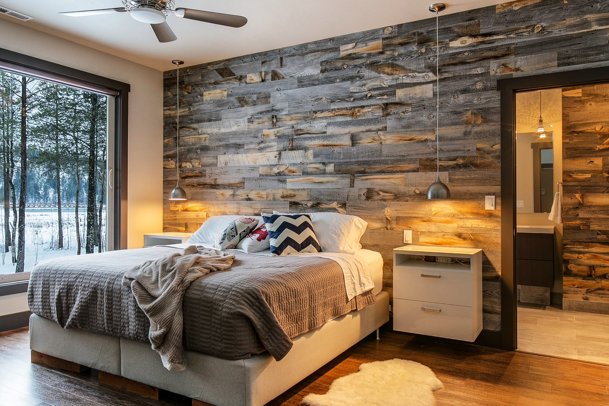 Cozy bedroom with a large window showing snow-covered trees outside, a wooden accent wall, a bed with throw pillows and blankets, two pendant lights, a ceiling fan, nightstands, and a doorway leading to a bathroom.