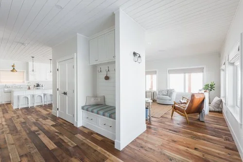 Open-plan living room and kitchen area with white walls, wooden flooring, and large windows with blinds.