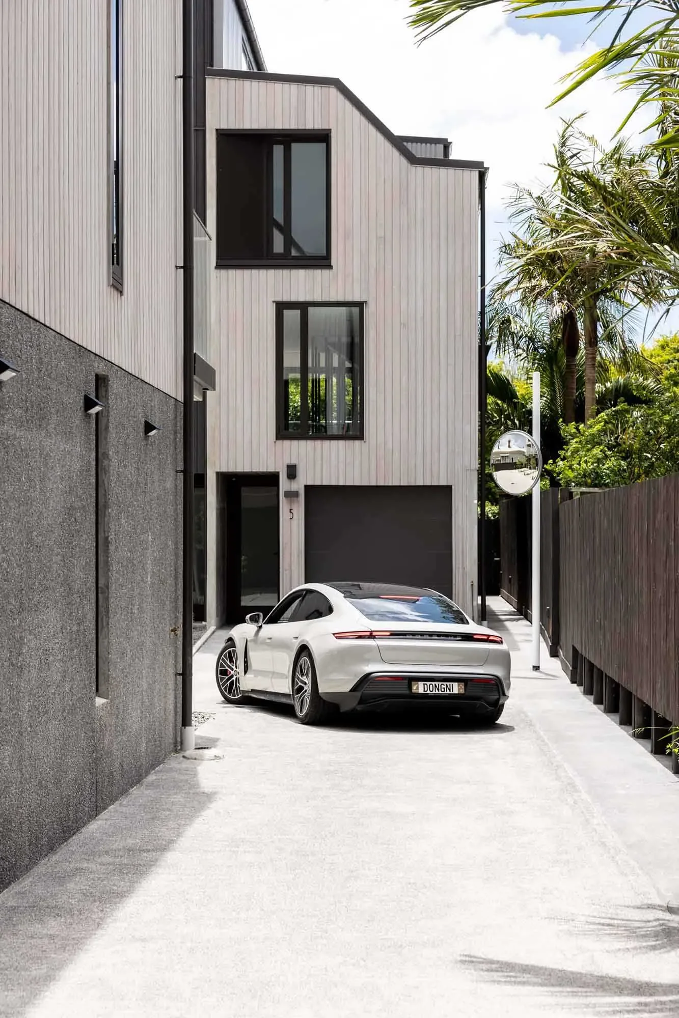 Modern house with black garage door and white luxury car parked in driveway, surrounded by palm trees and wooden fencing.