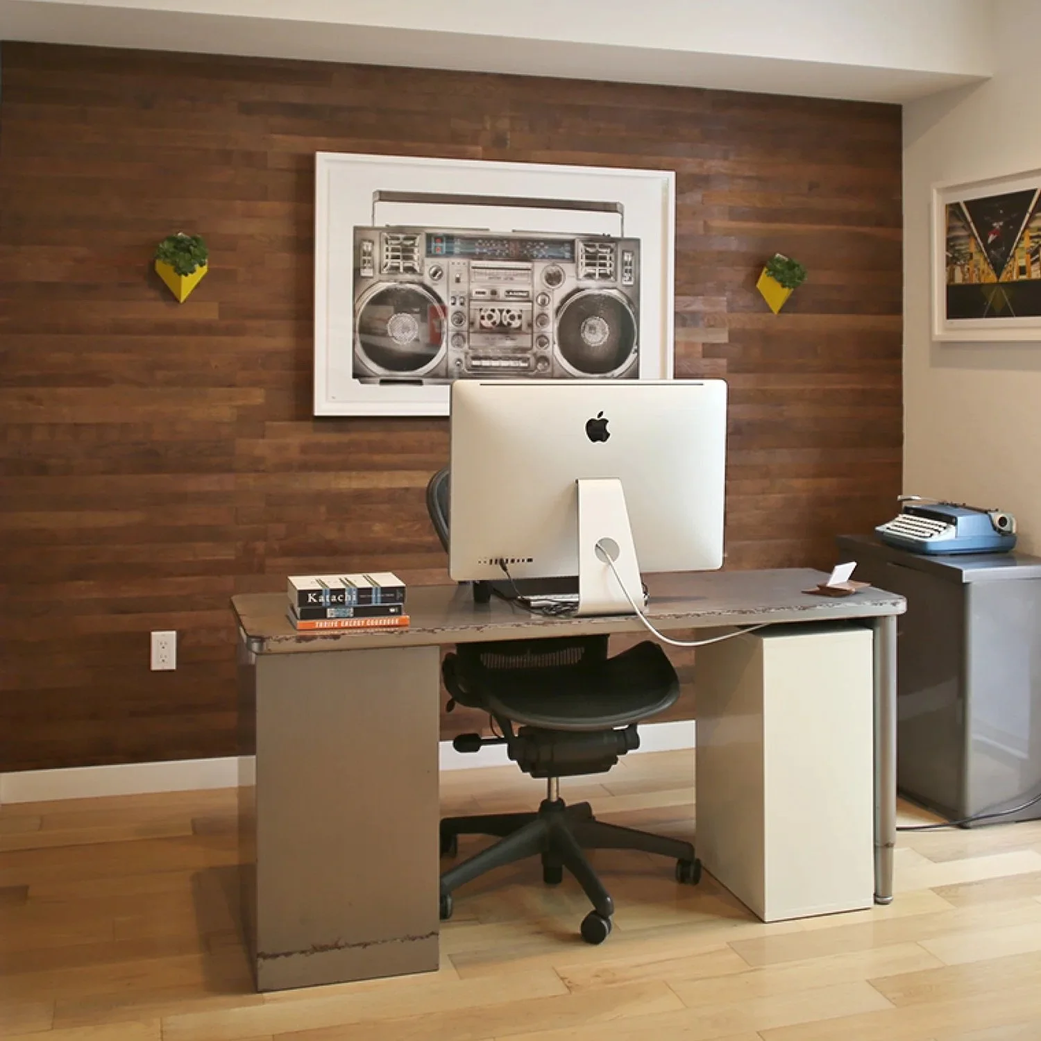 Office desk with an iMac computer, a stack of books, and a vintage typewriter, against a wooden paneled wall with framed artwork and plant decorations.