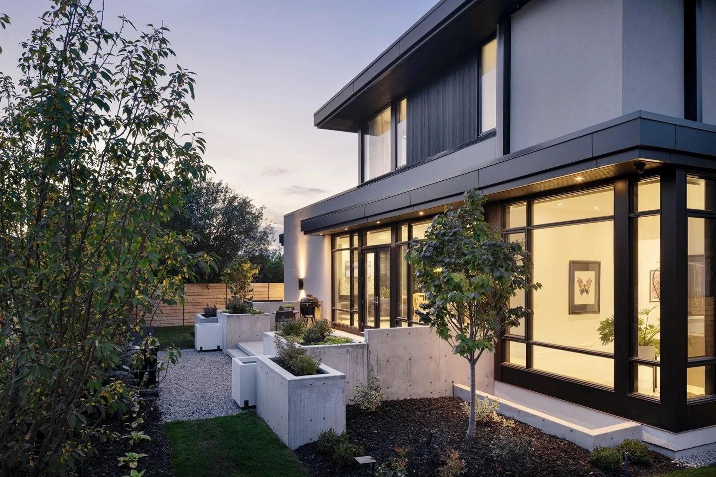 Modern two-story house with large glass windows and a landscaped backyard with trees and planters at dusk.