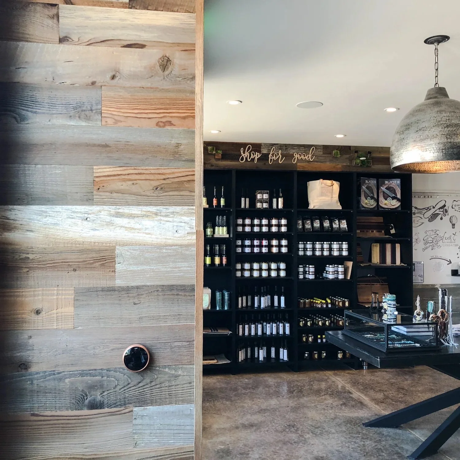 Interior of a retail shop with black shelving filled with jars and bottles, a wooden sign reading 'shop for good' above the shelves, a metallic hanging light, and a wooden wall on the left side.