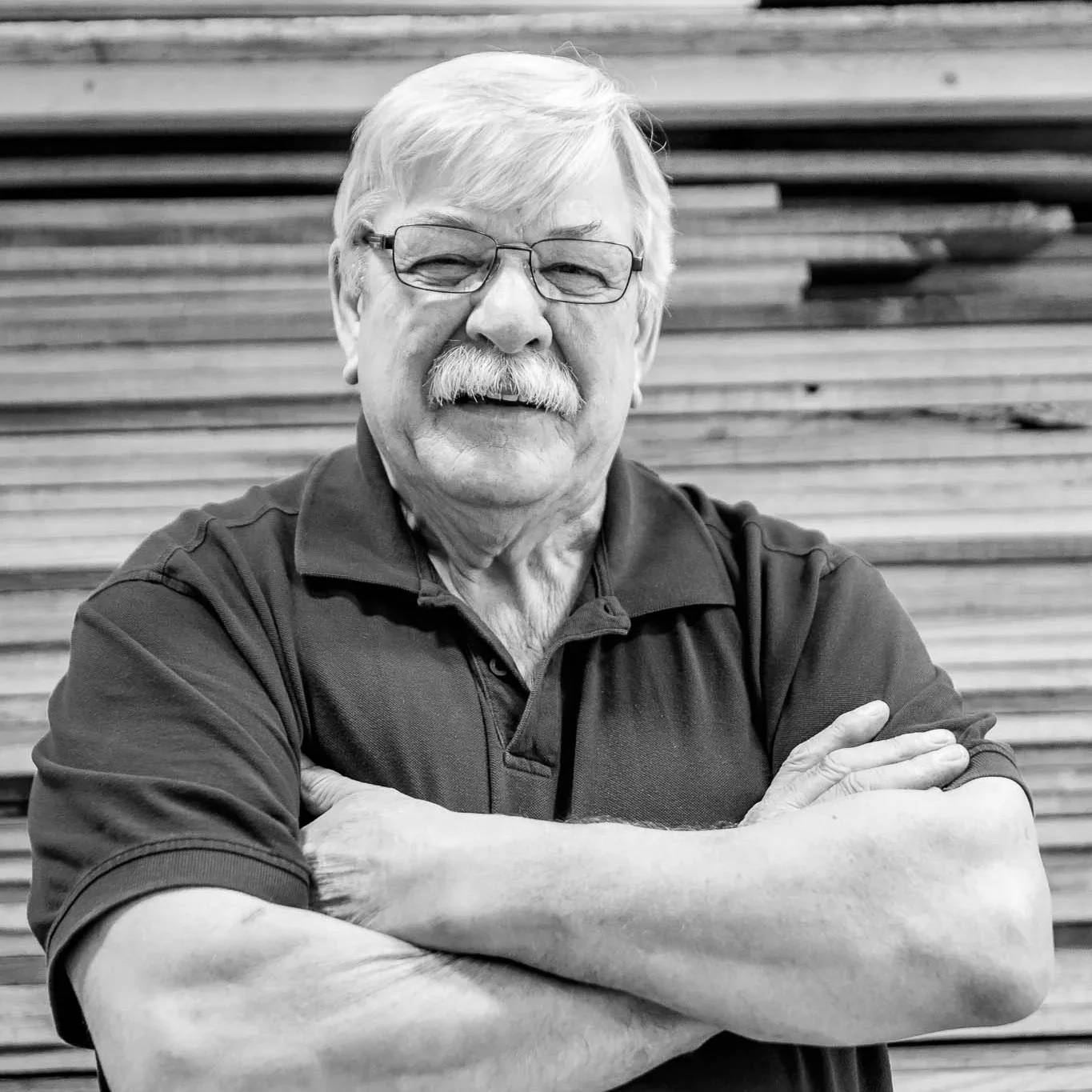An older man with glasses and a mustache, smiling with arms crossed, standing in front of a wooden wall.
