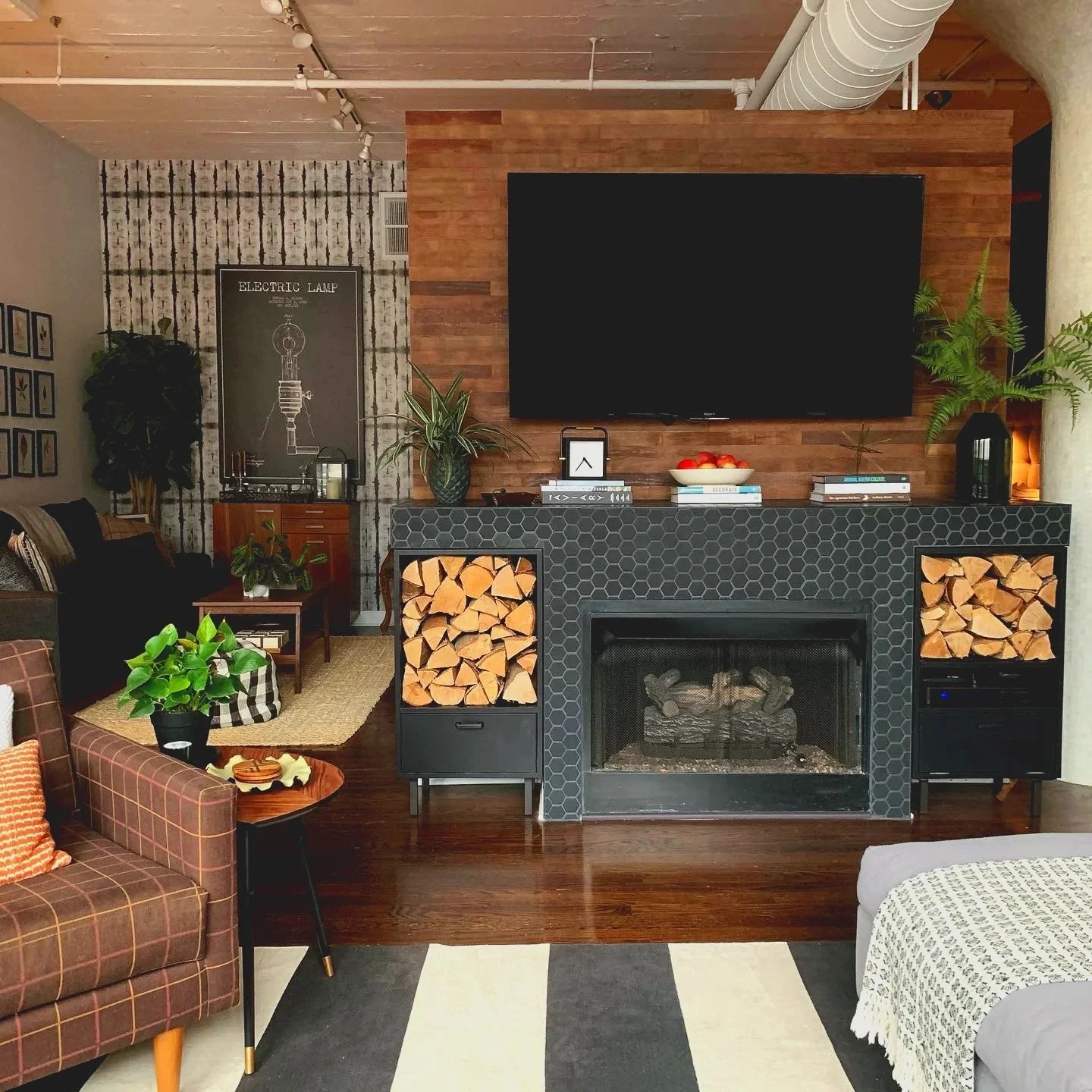 Living room with a fireplace, mounted flat-screen TV, wood logs, plants, and cozy furniture including a plaid armchair and a gray armchair with a blanket.
