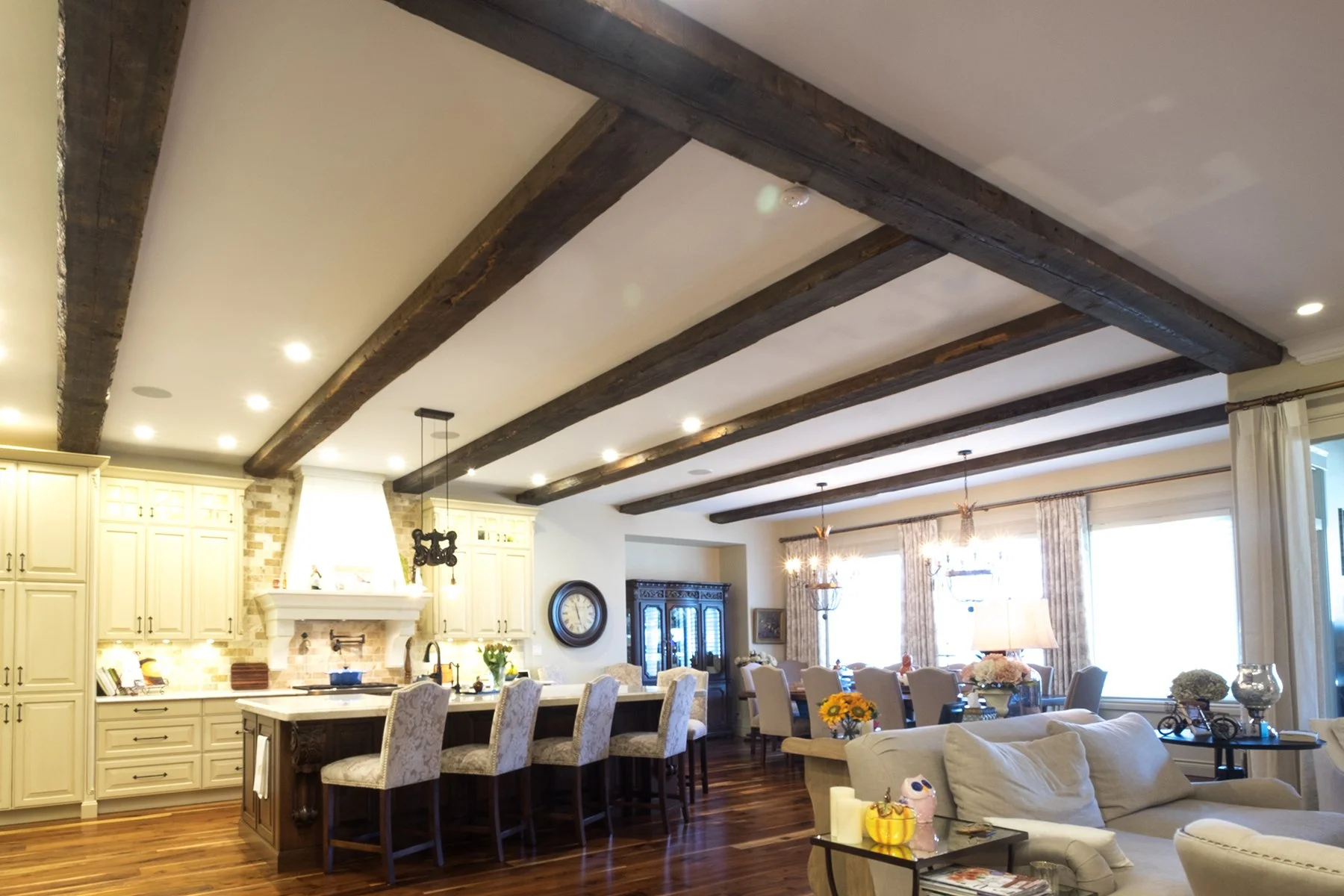 Open-concept living room and kitchen with exposed wooden beam ceilings, cream cabinetry, and large windows letting in natural light.