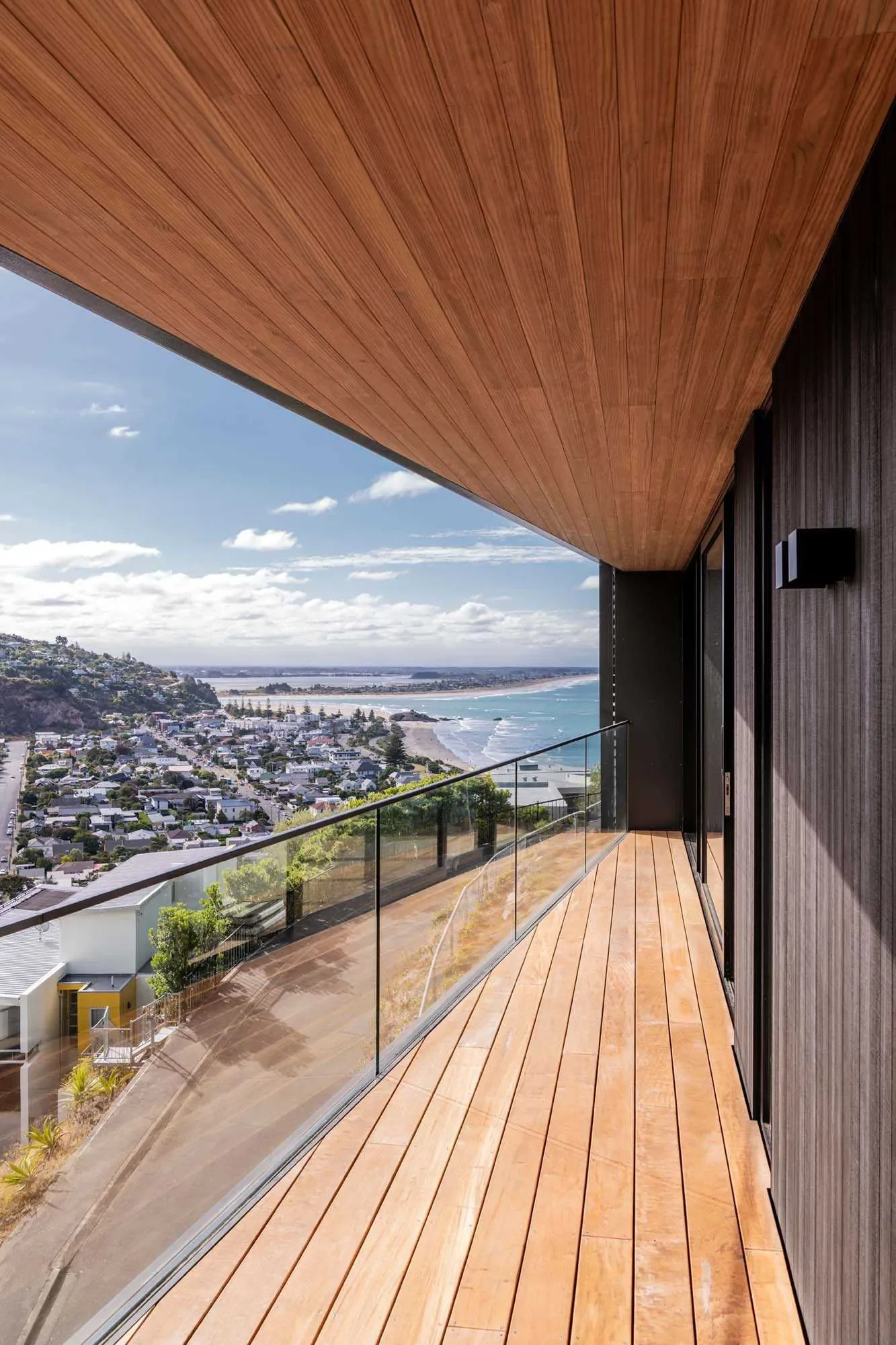 View from a modern balcony with wooden flooring and ceiling overlooking a coastal town and ocean.