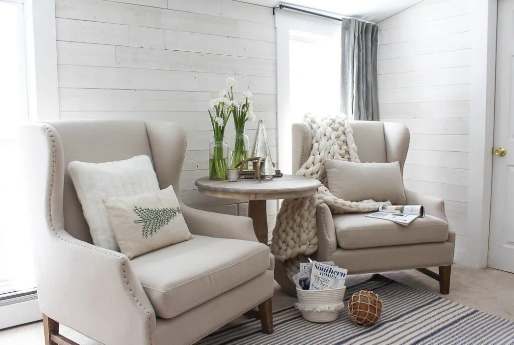 Cozy living room with two beige armchairs, a small round wooden table with vases of white flowers, pillows, a chunky knit blanket, magazines, and decorative items, with white wood-paneled walls and a window with curtains.