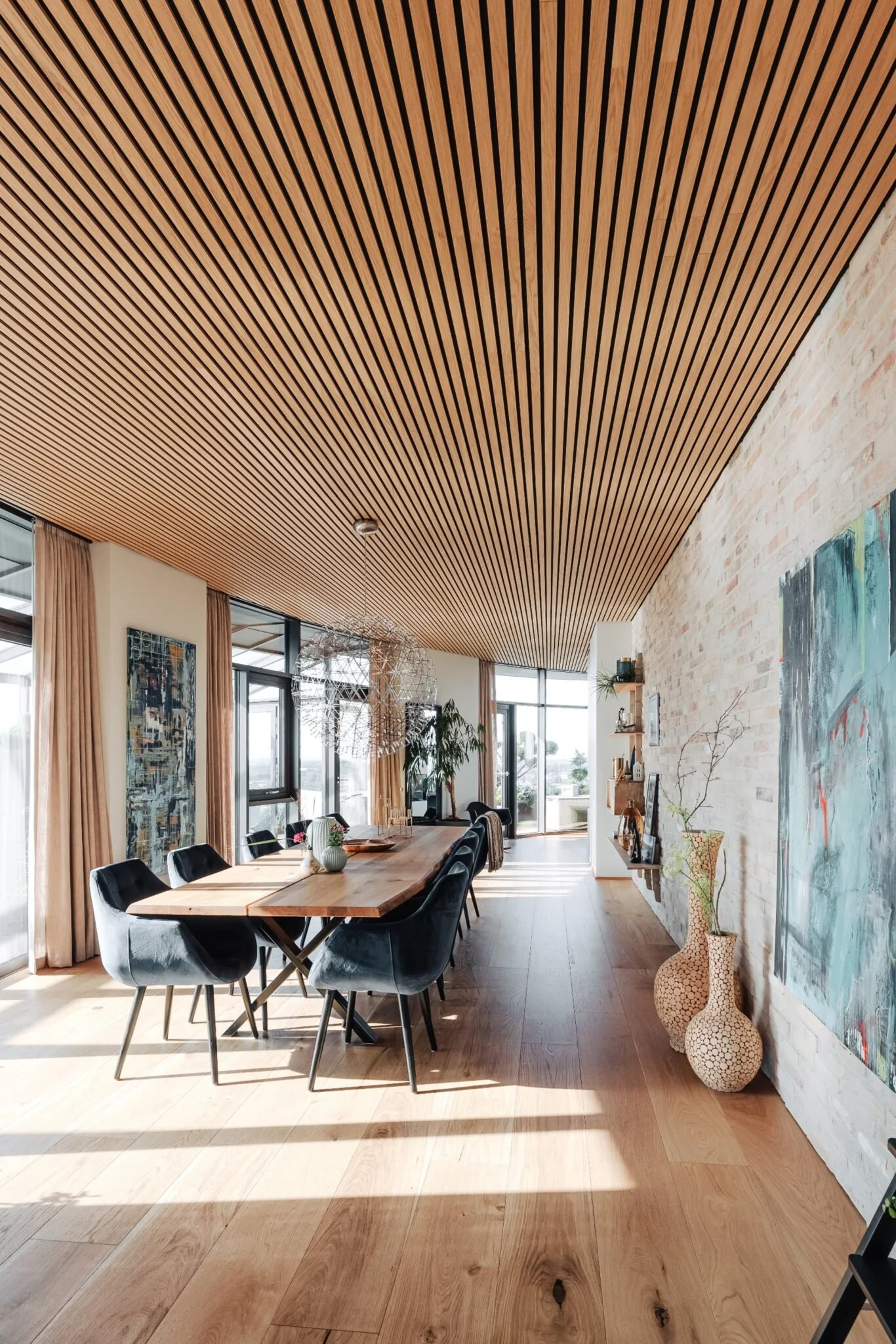 A modern dining room with a long wooden table, black chairs, large windows, a wooden slat ceiling, and a white brick wall decorated with artwork and vases.