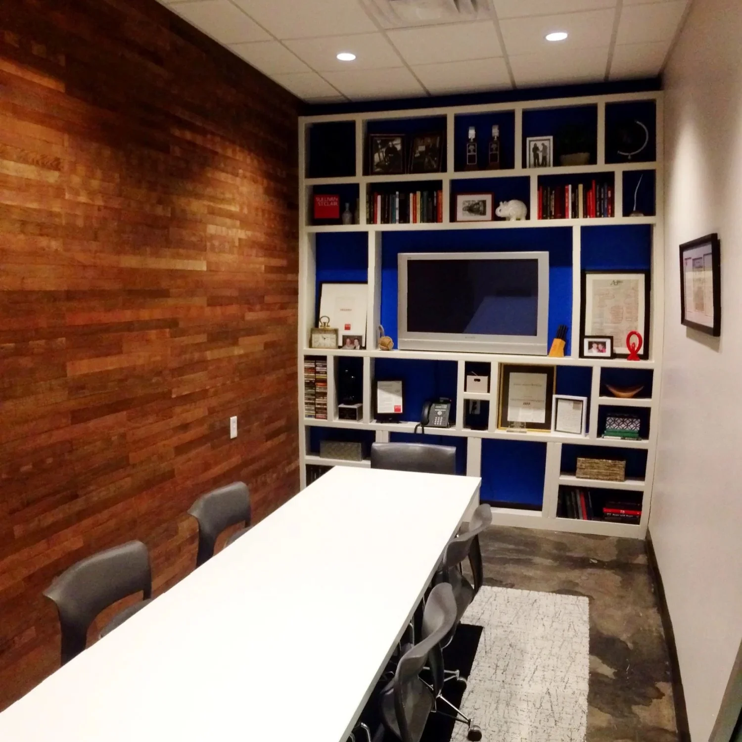 Conference room with a white table, six gray chairs, and a built-in bookshelf featuring a TV, books, framed photos, and decorative items against a painted wall.