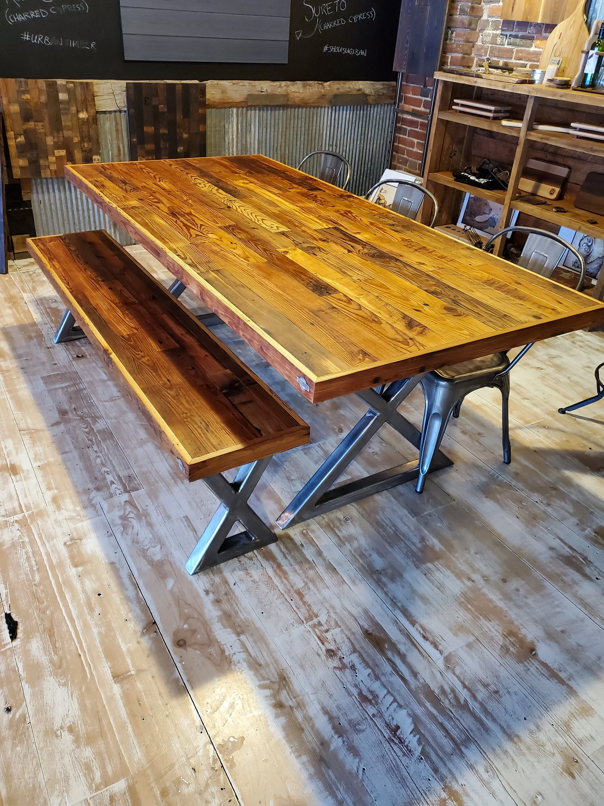 Wooden dining table with matching bench and surrounding metal chairs in a rustic restaurant with wood and brick decor.