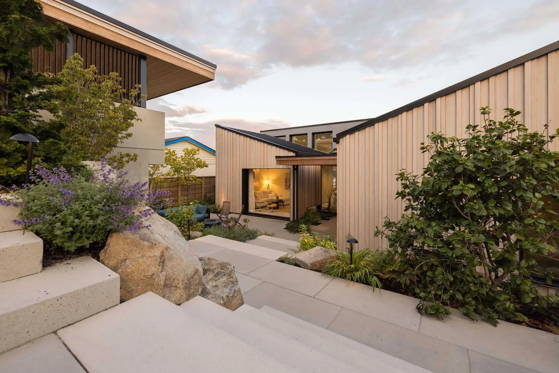 Modern house with wooden siding, outdoor seating area, landscaped garden, and stone steps during sunset