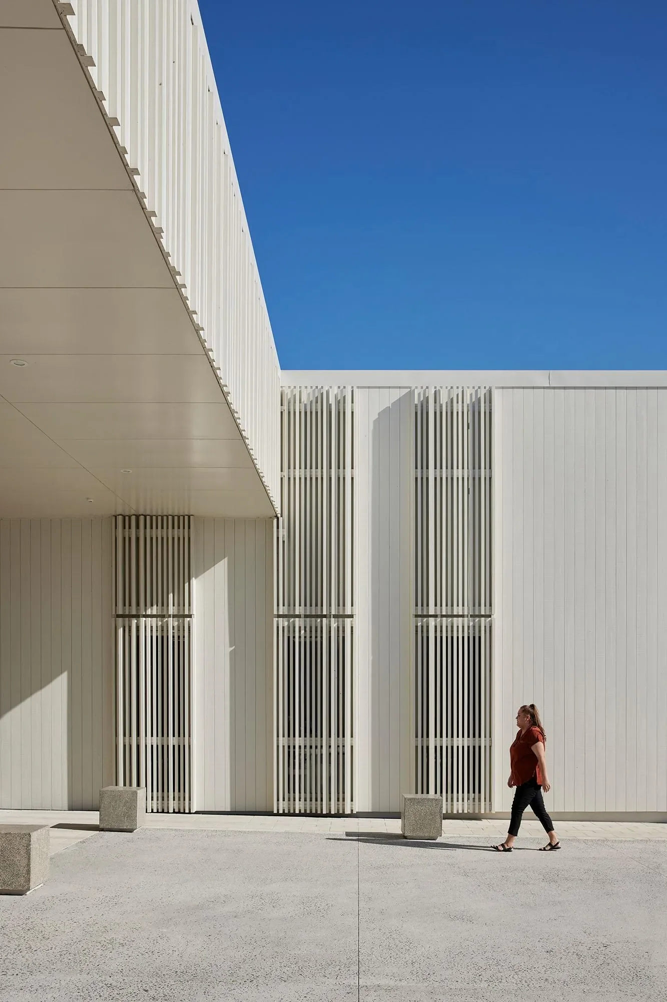 A woman walking past a modern white building with vertical slats and clean architectural lines under a clear blue sky.
