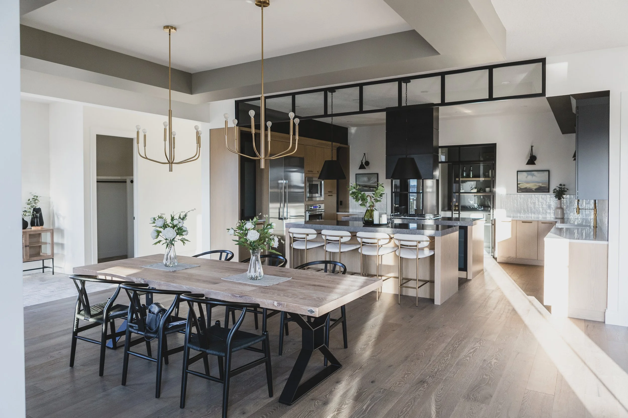 Open-concept kitchen and dining area with wooden table, black chairs, pendant lights, and barstools at a kitchen island. Decor includes glass vases with flowers.