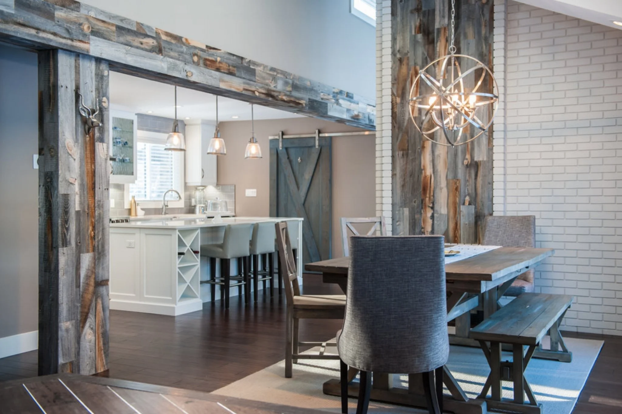 Interior view of a modern dining area with a rustic touch, featuring a wooden dining table, upholstered chairs, a hanging light fixture, and a partial view of a kitchen with white cabinetry and pendant lights.