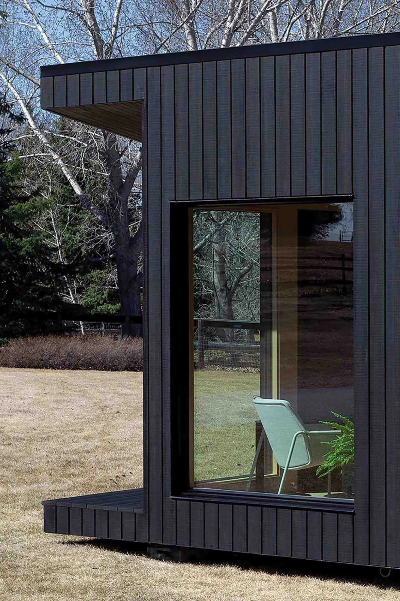 A modern black house with large glass windows and a small outdoor patio with a white chair and green potted plant, surrounded by a grassy yard and trees.