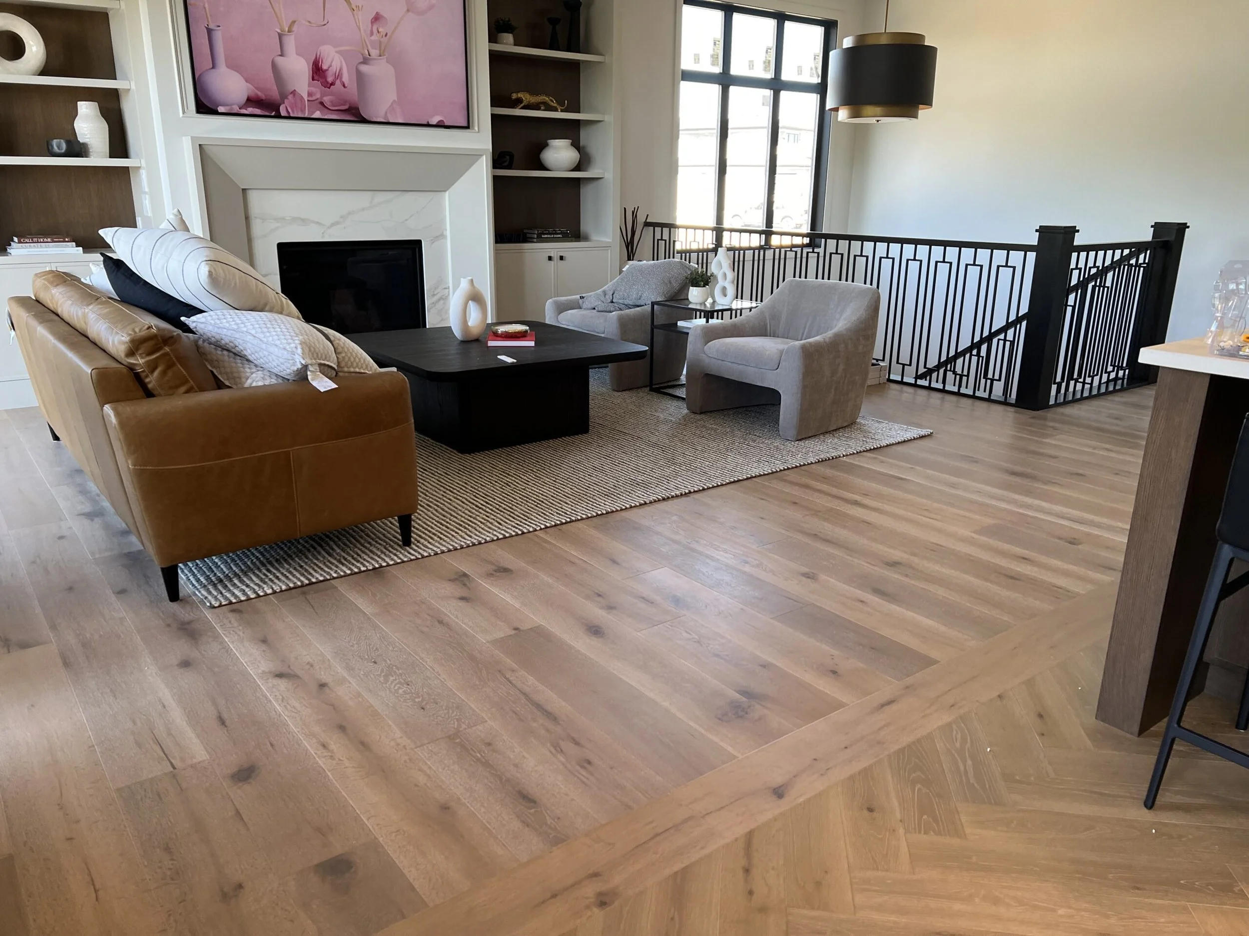 Living room with wooden floor, tan leather sofa, gray armchair, black coffee table, built-in shelves with decorative objects, fireplace, large window, staircase with black railing, and wall art depicting pink vases.
