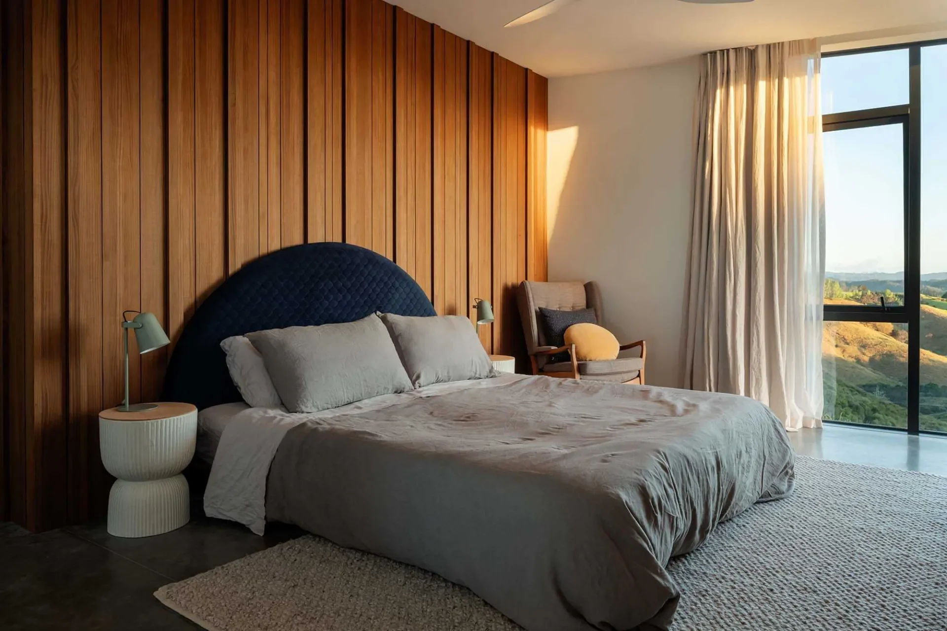 Modern bedroom with wooden paneled wall, large bed with pillows, a nightstand with lamp, armchair with cushions, large window with curtains showing scenic hills.