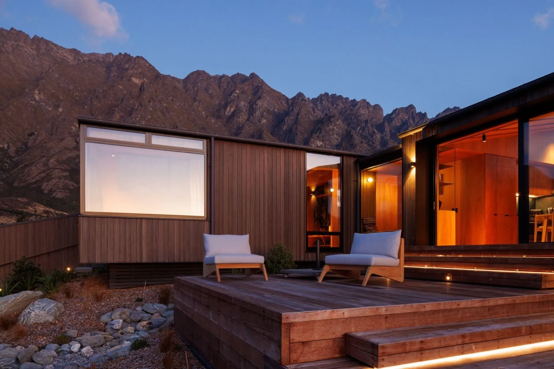 Modern wooden house with large windows and outdoor seating area set against mountain landscape at dusk.
