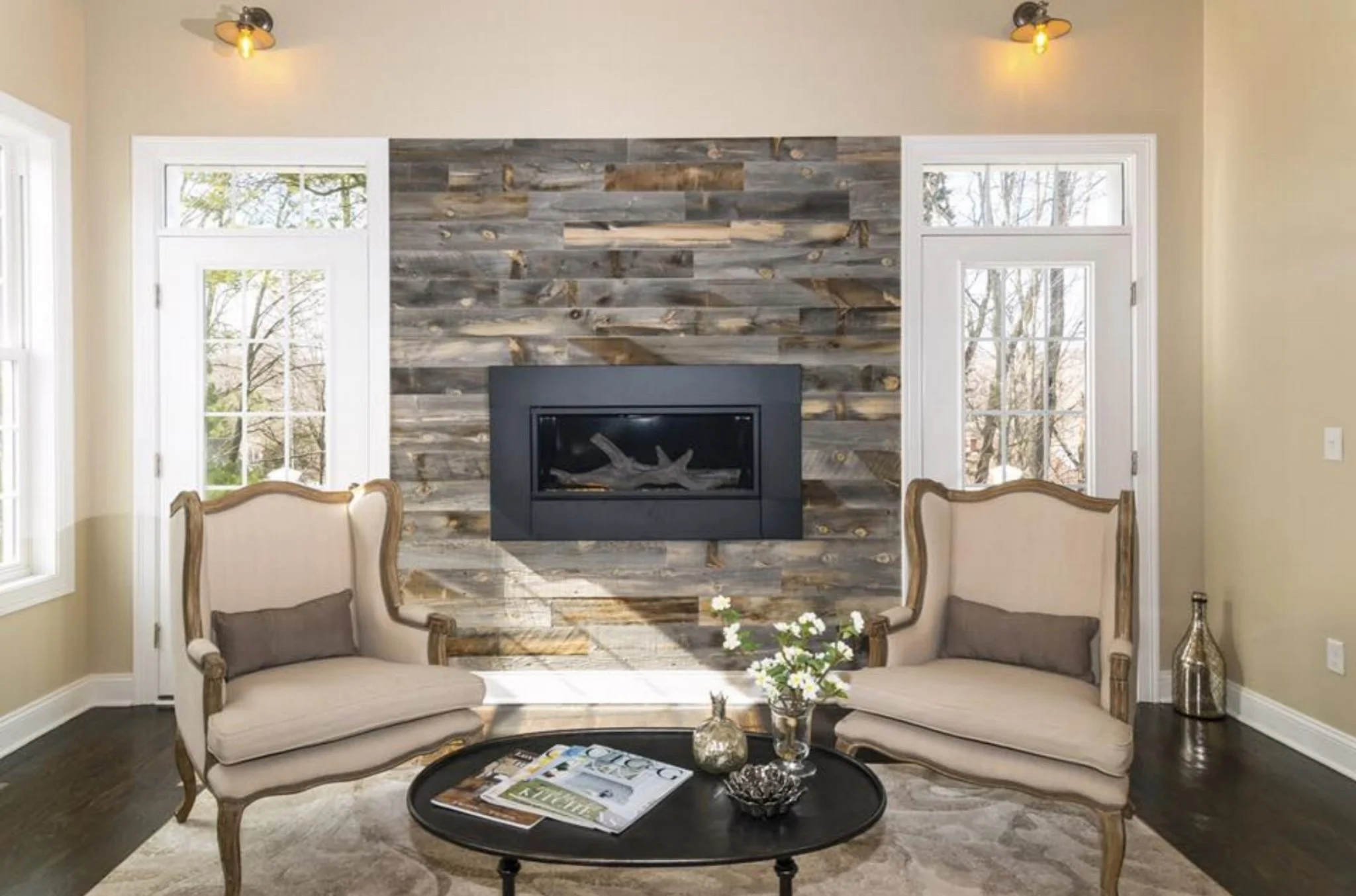 Living room with beige walls, two vintage armchairs with wooden frames and gray cushions, a black round coffee table with magazines, a glass vase with white flowers, and a decorative bowl. The back wall features a wood plank accent wall with a built-