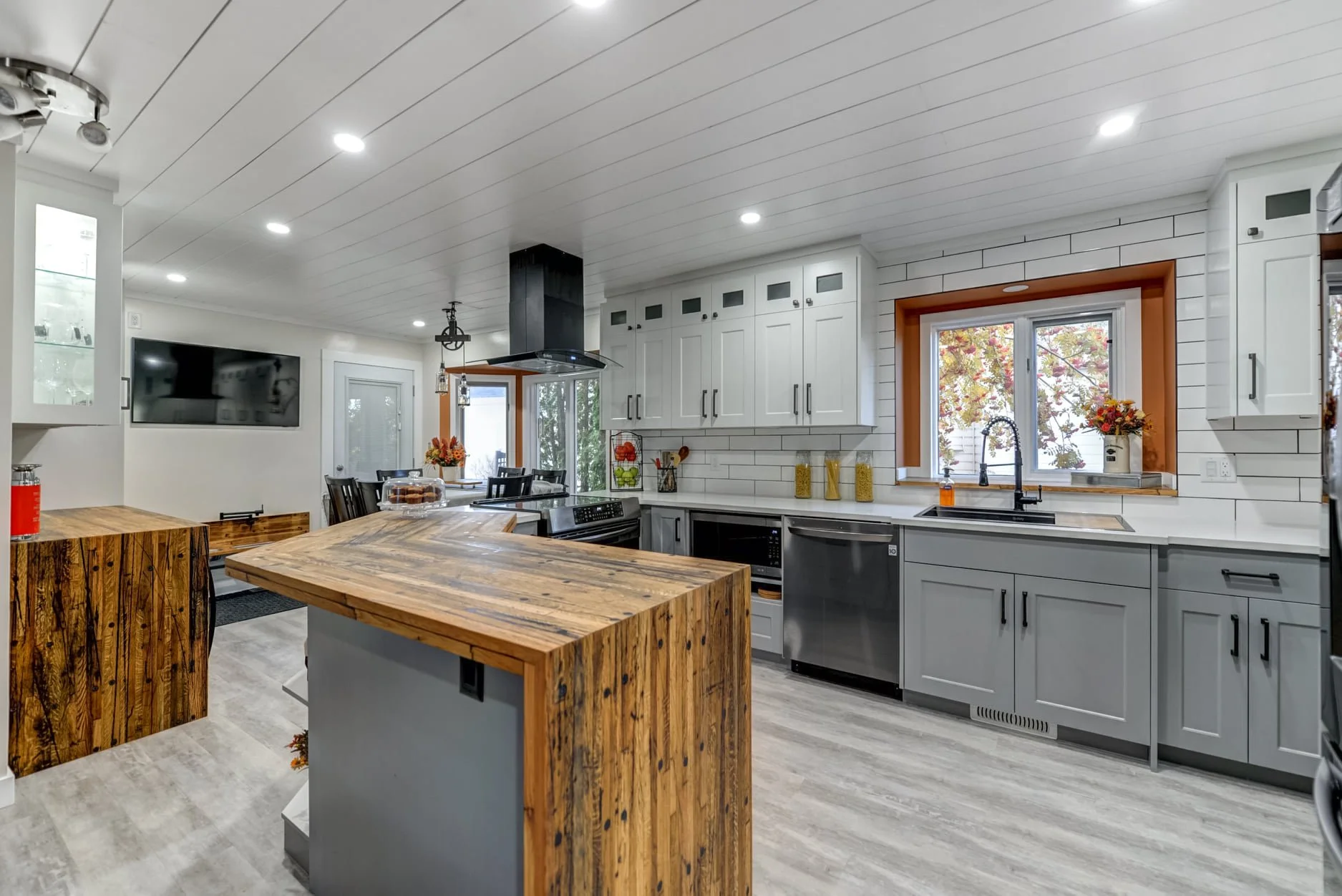 Modern kitchen with white cabinets, gray lower cabinets, and a wooden island. Includes a stainless steel dishwasher, microwave, and black range hood. Features a window with a view of trees with fall foliage and decorative items like flowers and jars.