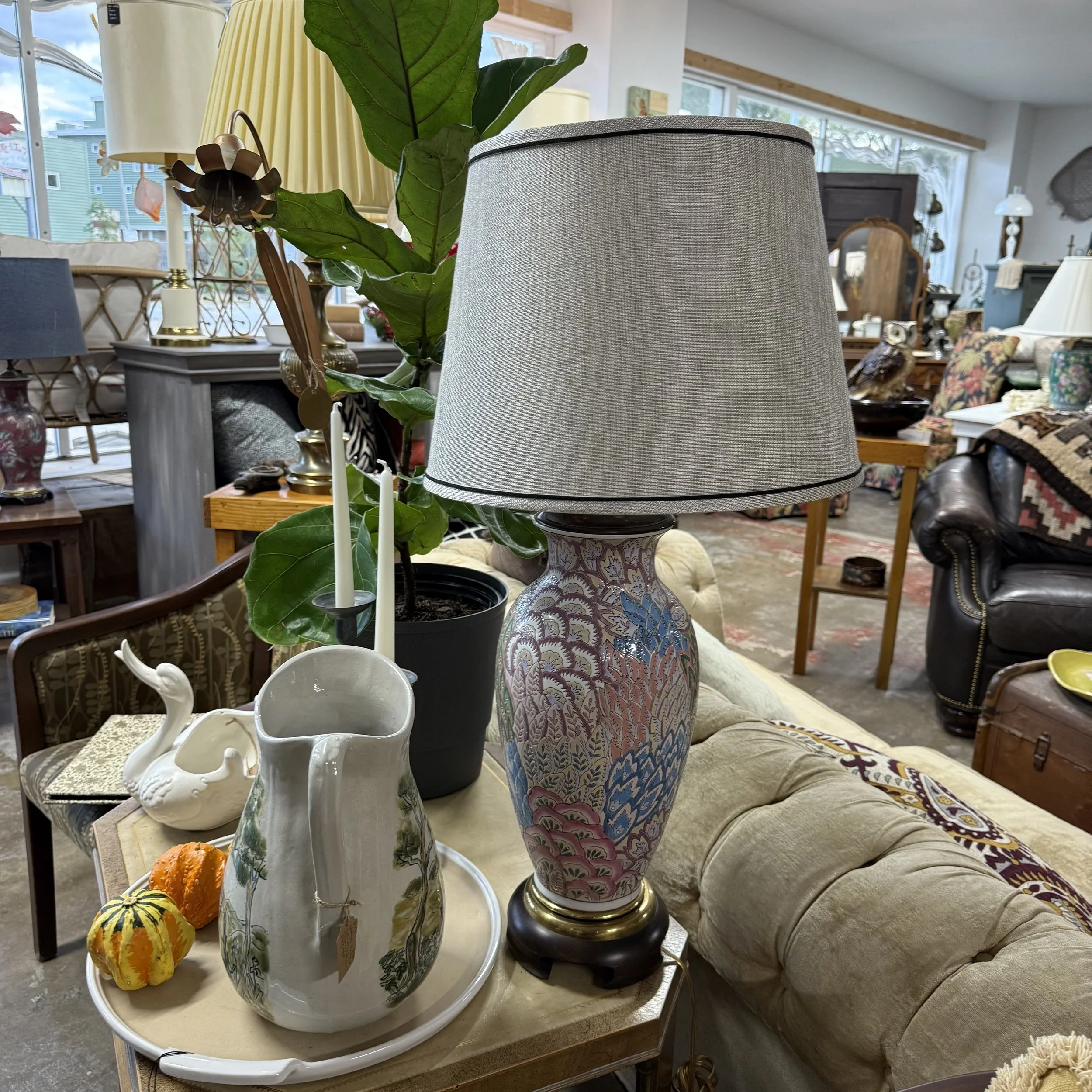 Furniture and decorative items in an indoor space, including a table with a large ornate table lamp, a white swan-shaped ceramic pitcher, a white ceramic pitcher with a landscape design, small decorative gourds, and a large potted leafy plant, surrou