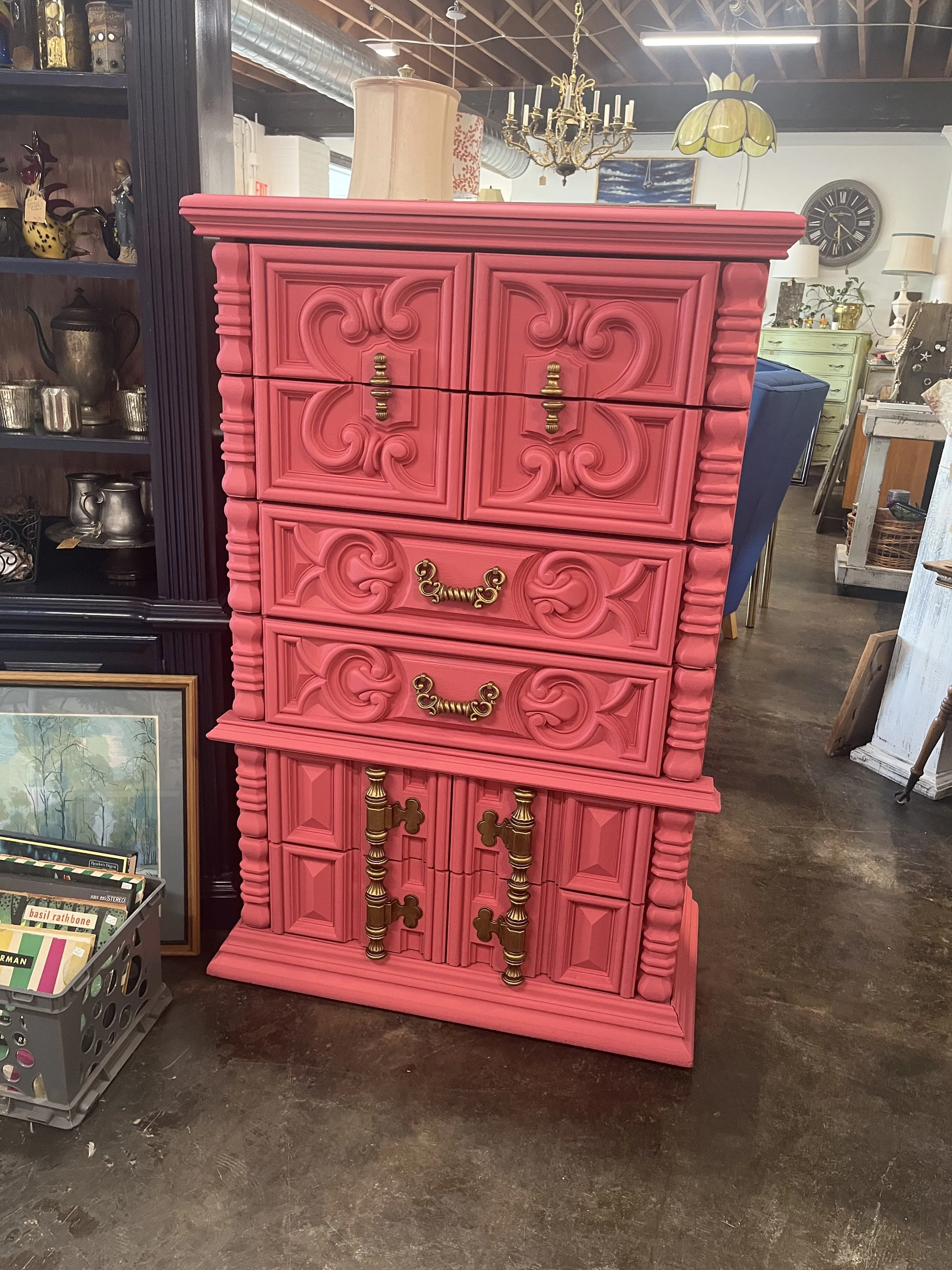 A bright pink vintage-style dresser with ornate carvings, gold handles, and multiple drawers, placed in an antique store.