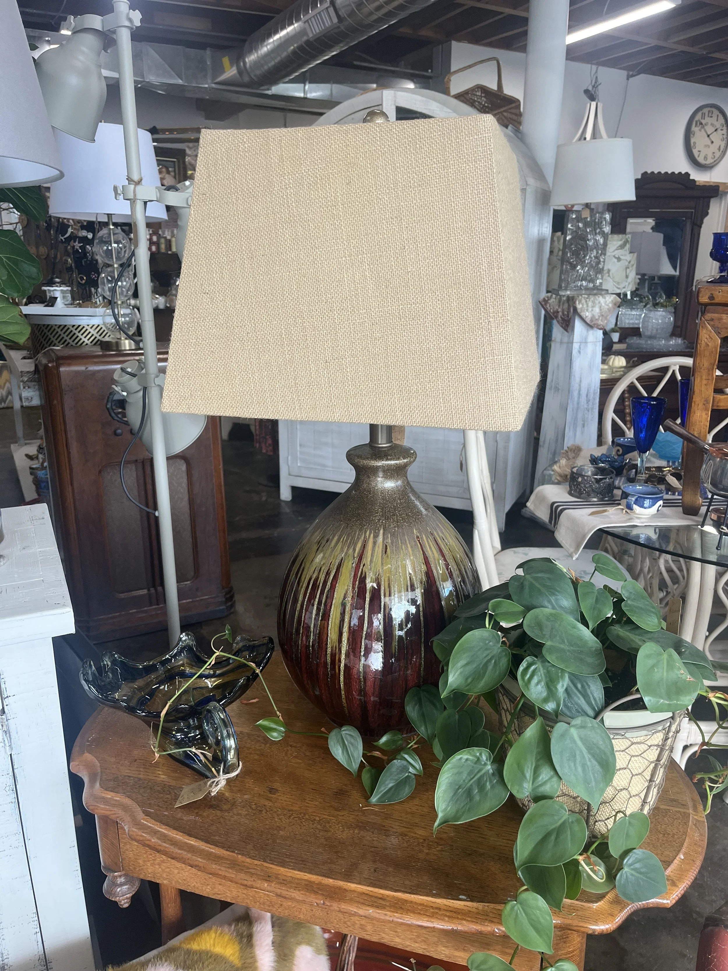 A large ceramic table lamp with a beige lampshade, placed on a wooden table alongside a green potted plant and a decorative glass bowl in an antique or thrift store setting.