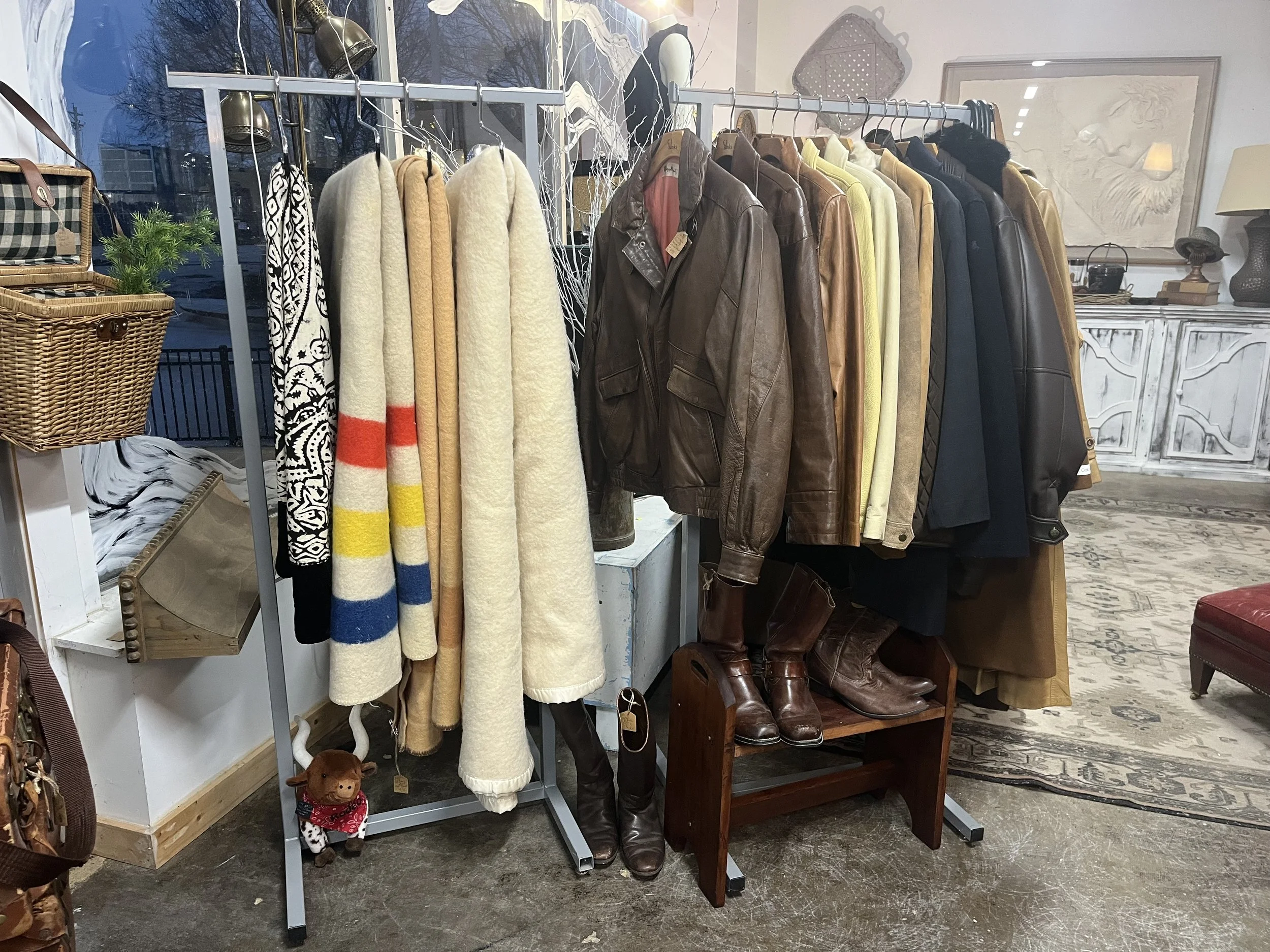 A clothing rack with various coats and jackets, including leather and wool options, placed inside a store. A pair of brown leather boots is on a small stand beneath the jackets. To the left, there are scarves, hats, and a small stuffed dog with a red bandana sitting on the floor. The background shows a decorated interior with framed artwork, a white cabinet, and a rug.
