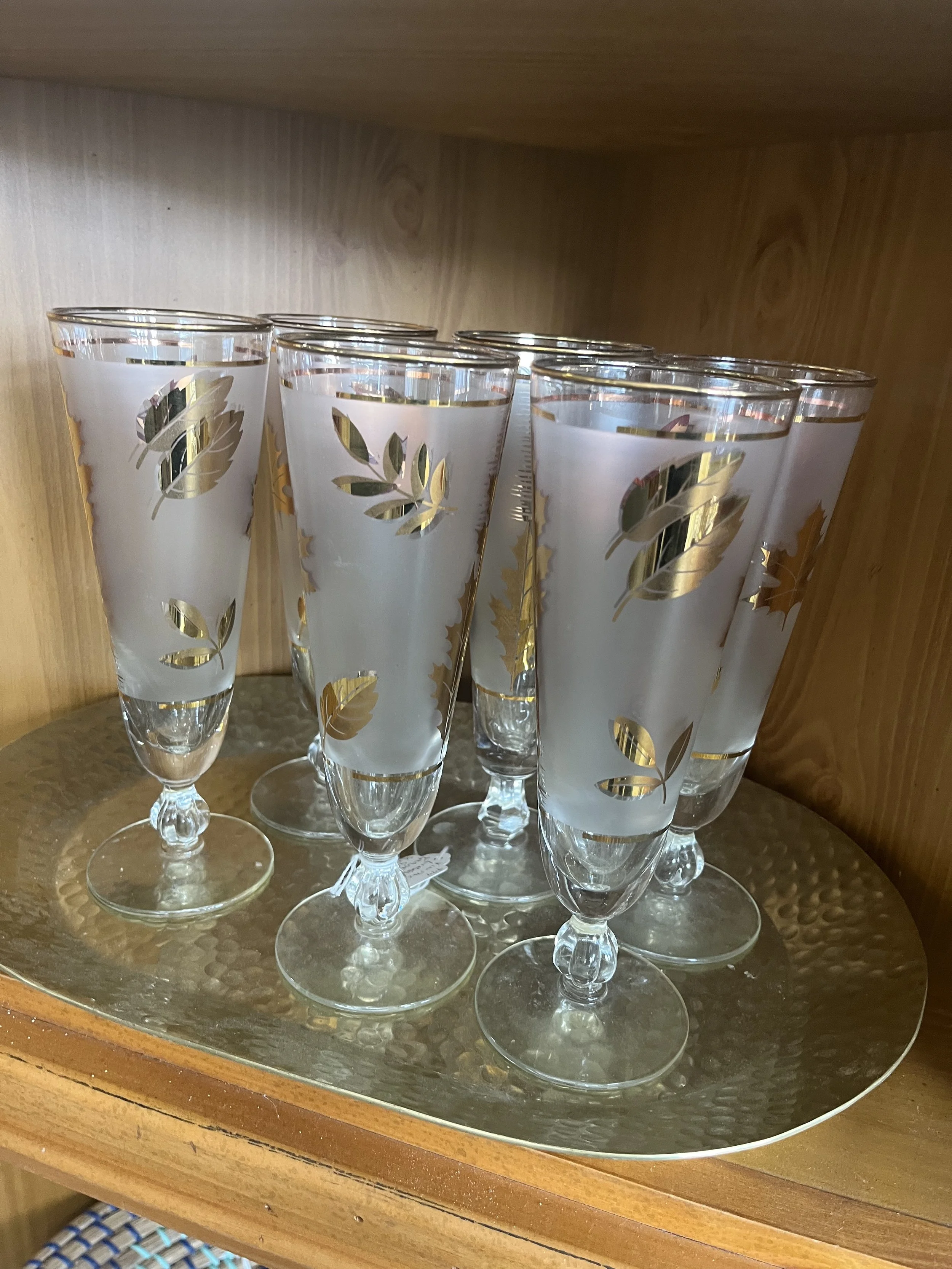 Set of frosted champagne glasses with gold leaf design on a textured gold tray inside a wooden cabinet.