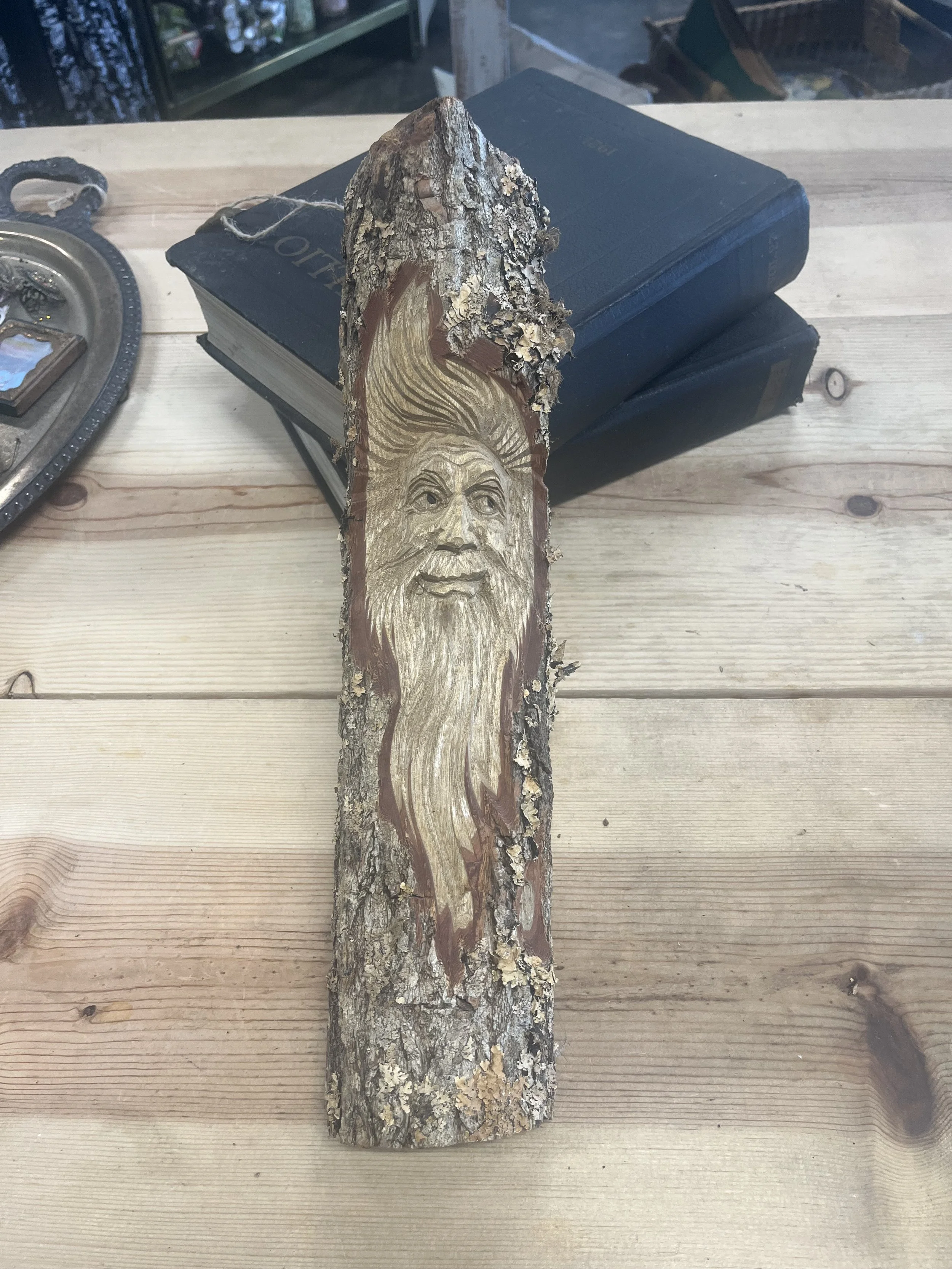 Decorative wooden piece with carved and painted face of a bearded man on a bark slab, on a wooden table with books and decorative items in the background.