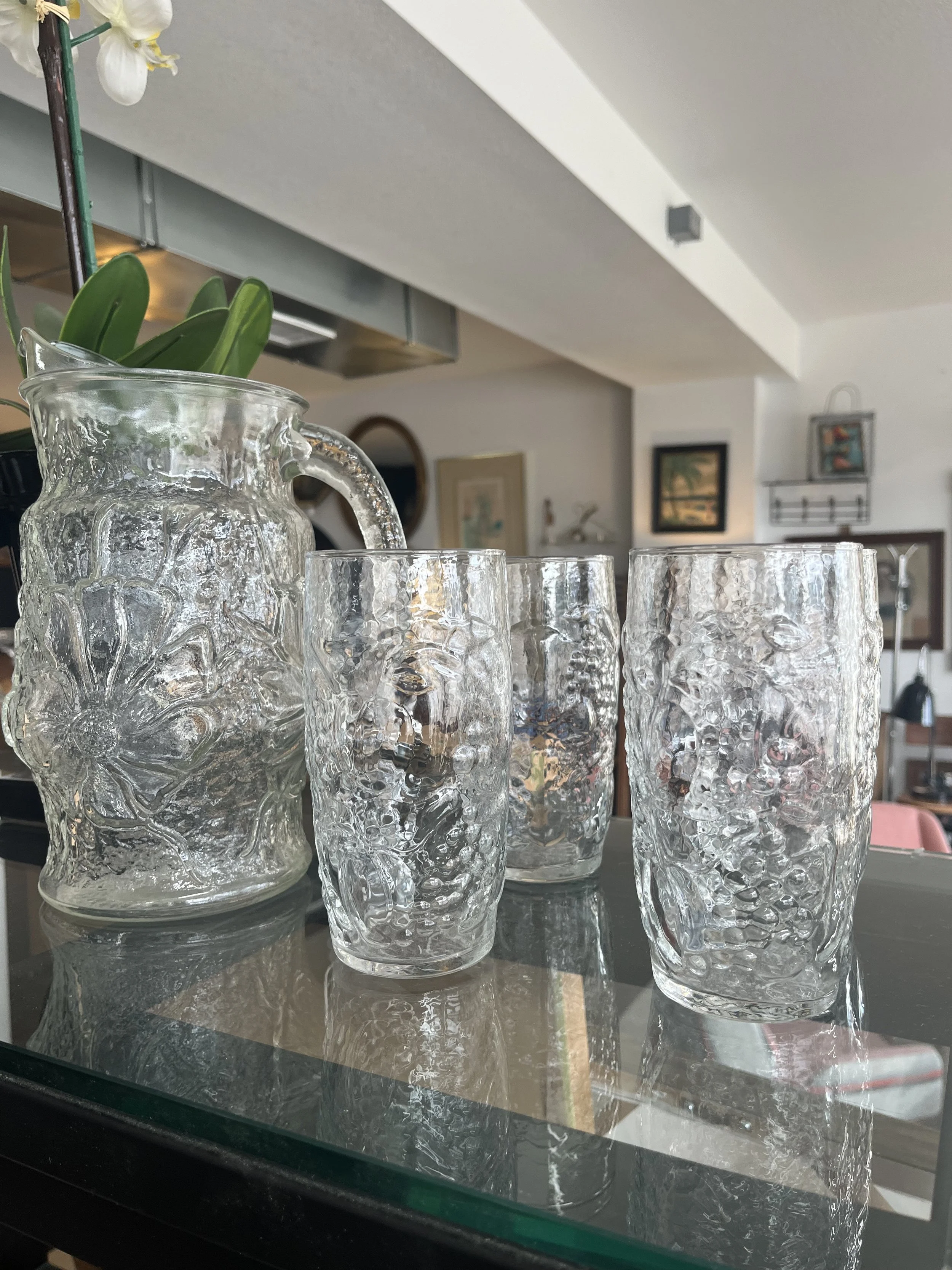 A decorative glass pitcher with an embossed floral design and three matching glasses on a reflective surface, with a background of a living room with framed pictures and plants.