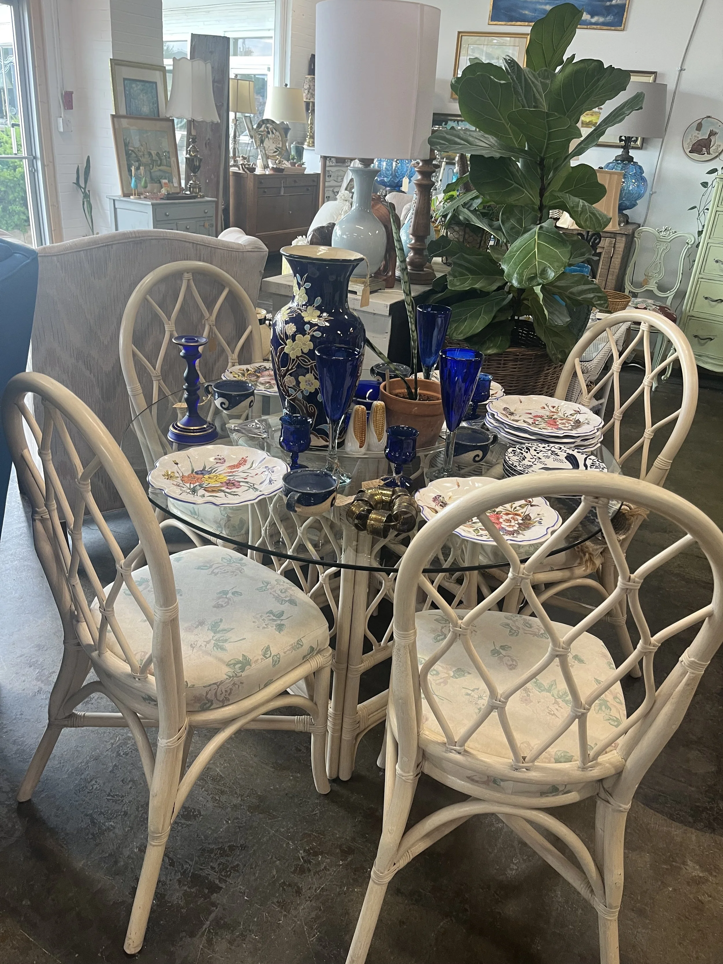 A round glass dining table set with floral dishware, animated by five white wicker chairs with floral cushions, decorations including blue vases, candles, and a potted large green plant in a woven basket, surrounded by various lamps and furniture in 