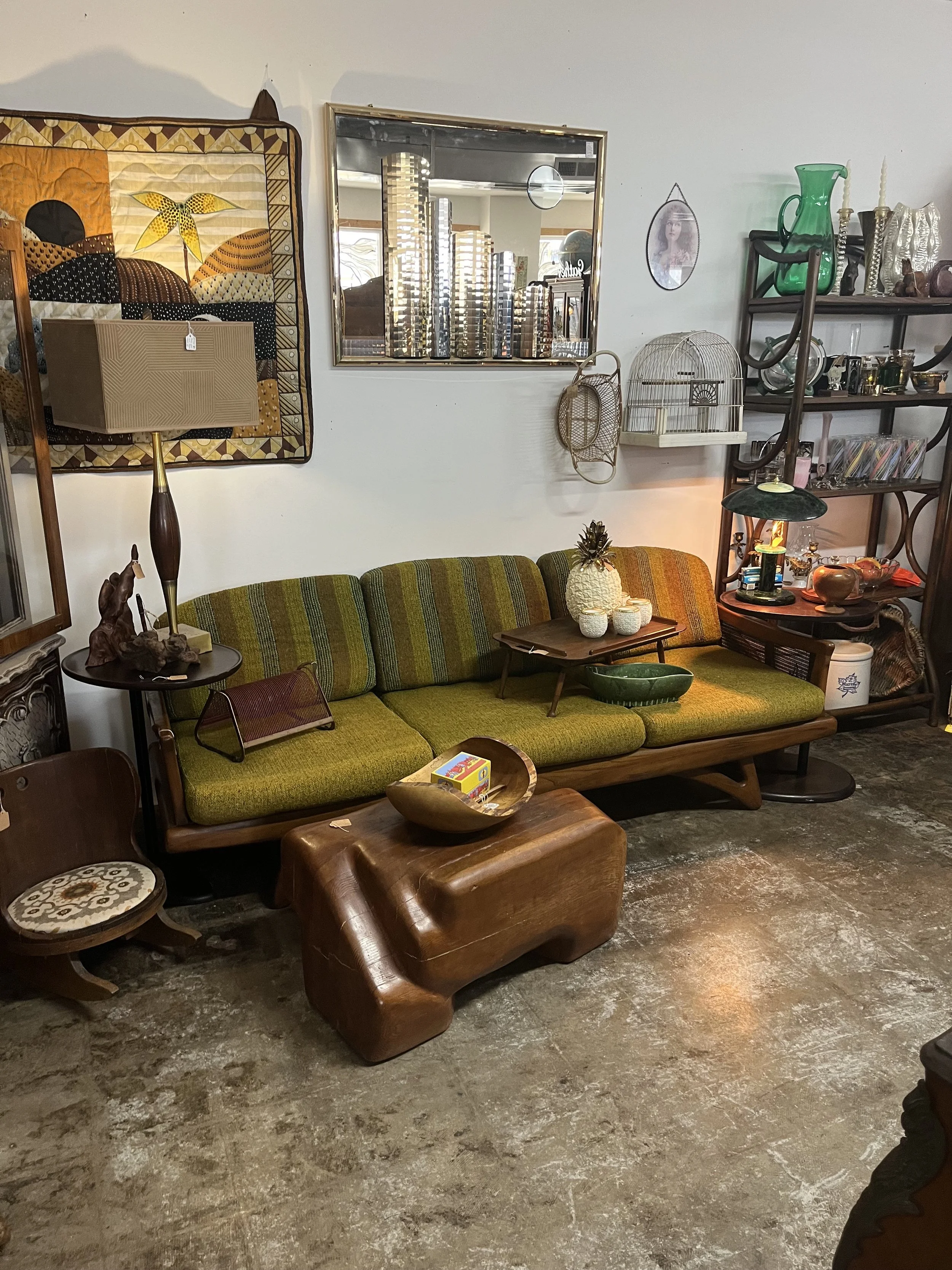 A vintage living room with a green striped sofa, a wooden coffee table, a small wooden chair, and various decorative items including a table lamp, framed artwork, and shelves filled with collectibles.