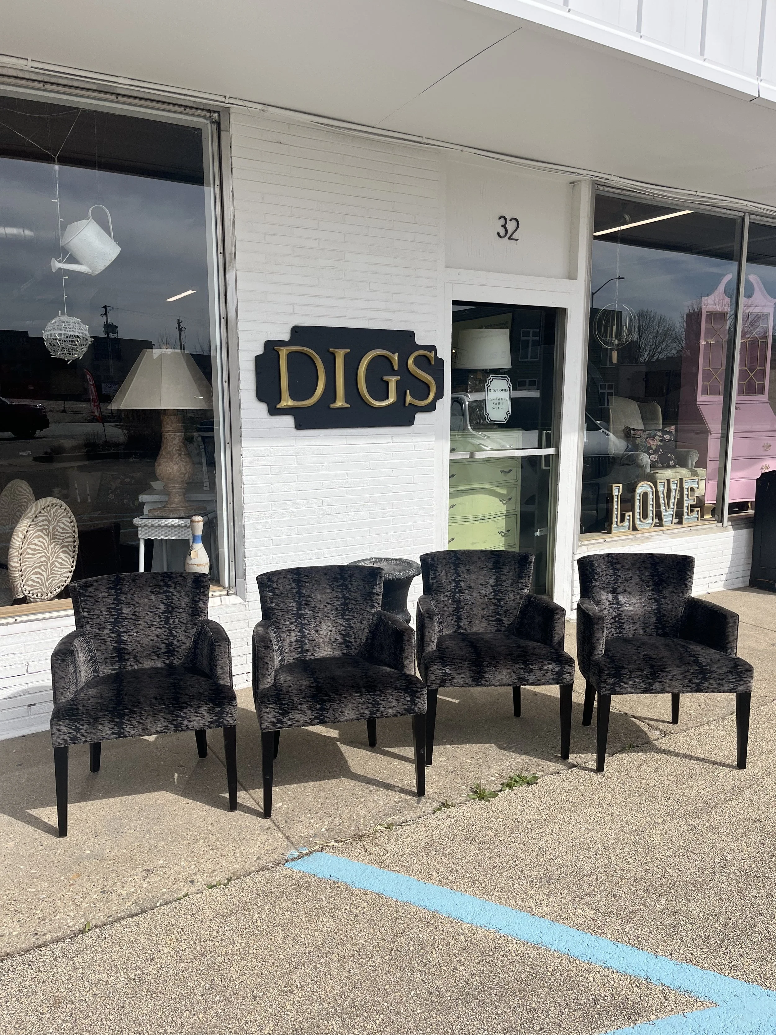 Four black upholstered chairs are arranged in front of a storefront with white brick walls. The store has a sign that reads 'DIGS' and displays decorative items in the window, including a lamp and the word 'LOVE' in large letters.