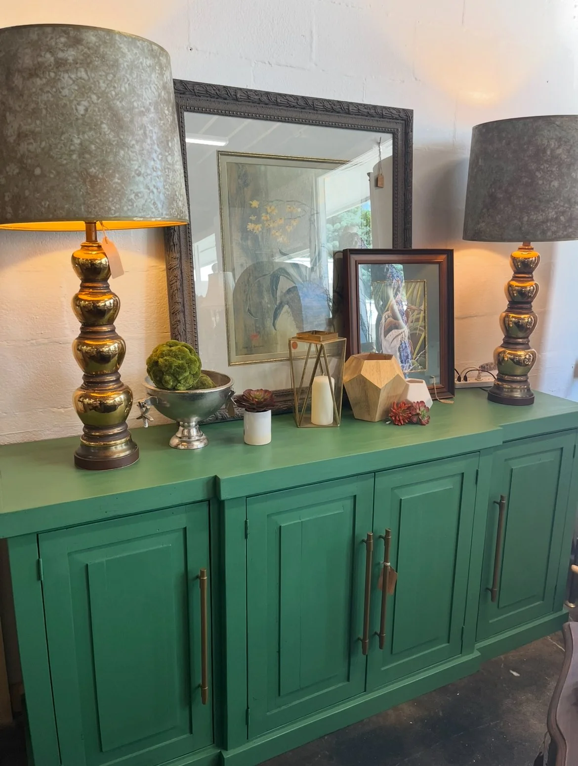 Green sideboard with decorative items, framed artwork, lamps, and plant decor.